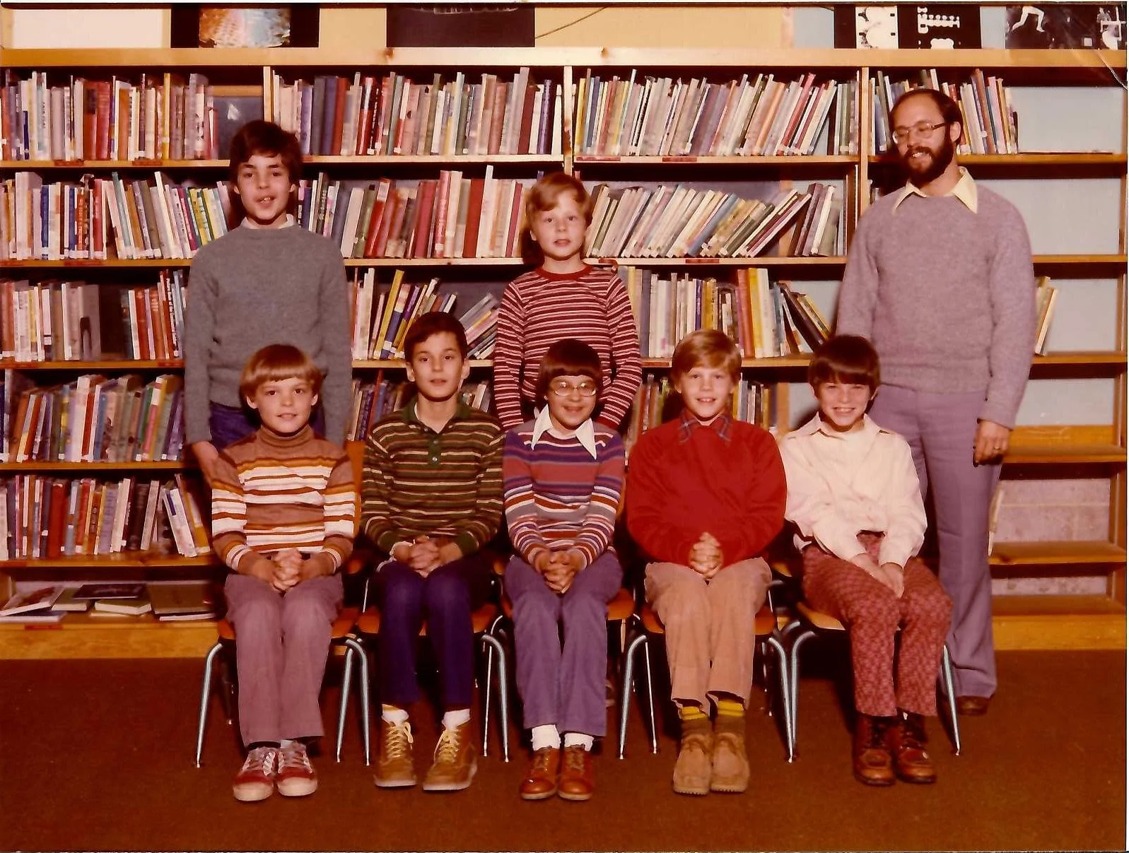 A group of eight children and one adult man in a library, sitting and standing in front of a bookshelf filled with books. The children are arranged in two rows with four children sitting in the front and three children standing behind, along with the man standing on the right side of the back row. The children are dressed in colorful casual clothing, and the adult man is wearing glasses and a sweater.