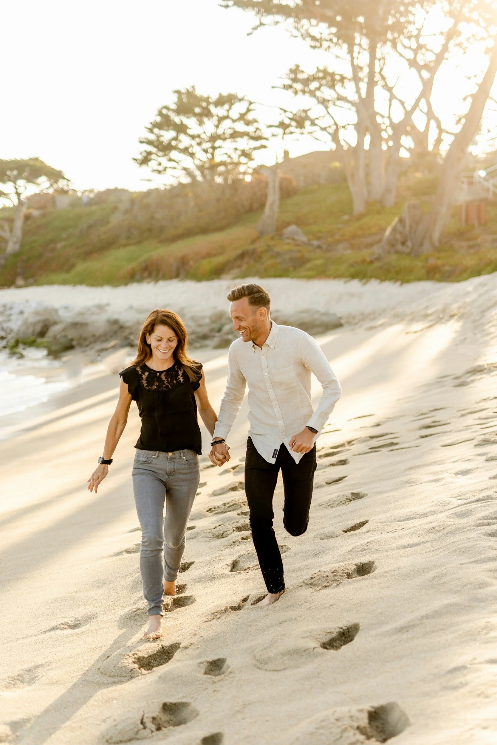 A young couple smiling and holding hands while walking barefoot on the beach, leaving footprints in the sand with trees and a hill in the background, satisfied with the sexual health services they receive from Wellness from Within in Knoxville, TN.
