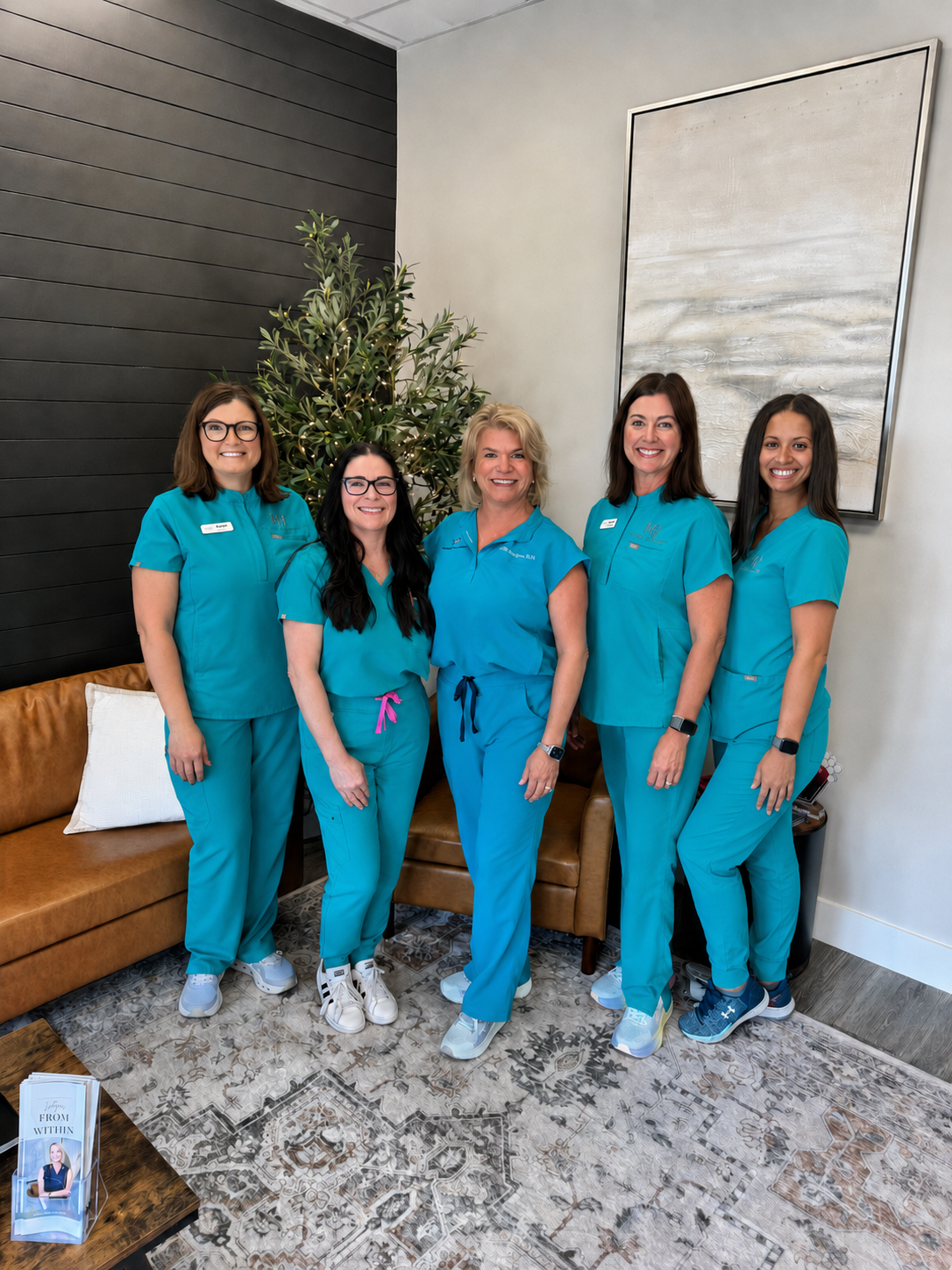 The amazing staff at Wellness from Within, in matching blue medical scrubs, standing their modern Knoxville office with a plant, a leather couch with a white pillow, and a large abstract painting on the wall.