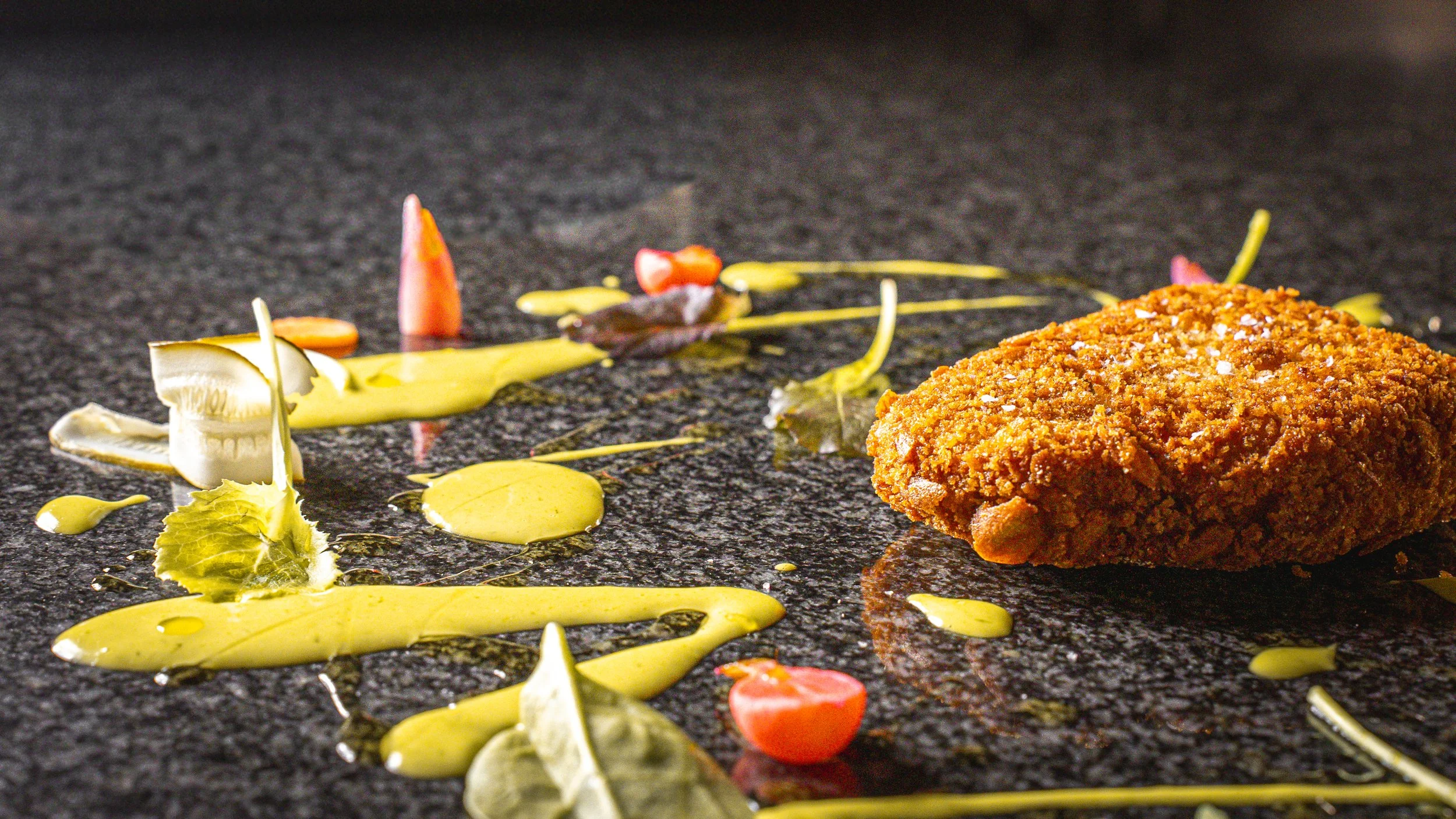 Frittura di pollo con decorazioni di salsa e verdure su superficie scura.