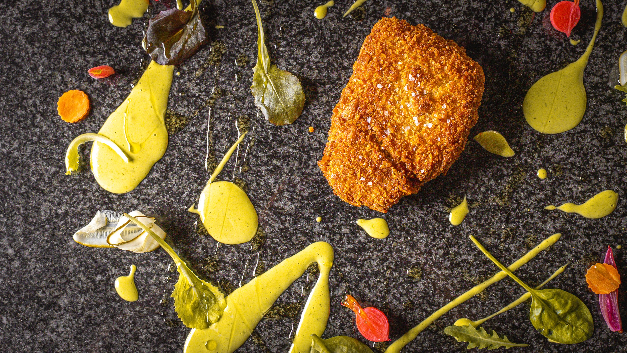 Un piatto gourmet con un pezzo di cibo impanato e fritto, decorato con diverse salse verdi e gialle, foglie e petali di fiori colorati, disposto su una superficie nera.