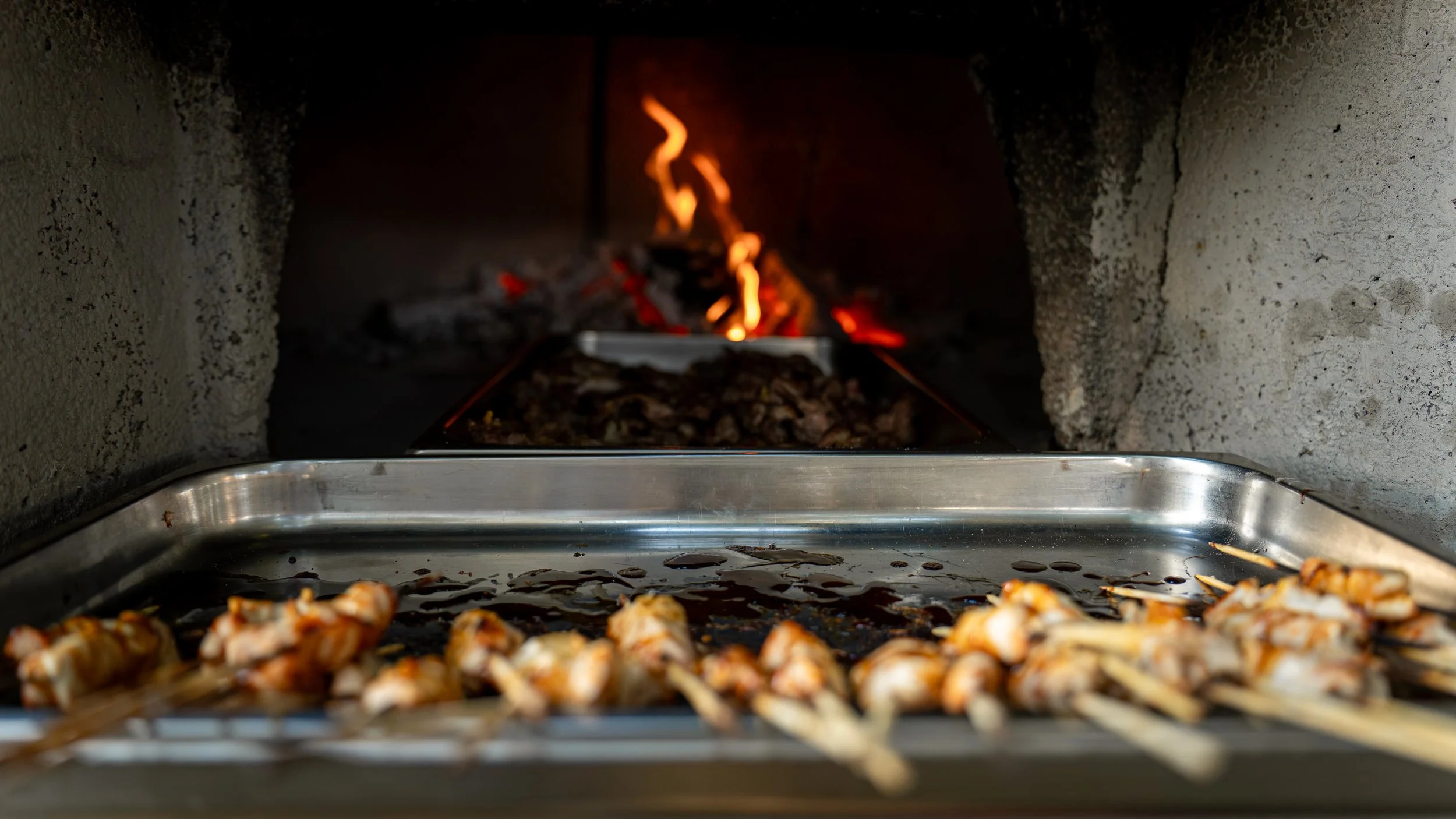 Cibo in cottura nel forno a legna, con una teglia di polpette e un fuoco acceso sul fondo