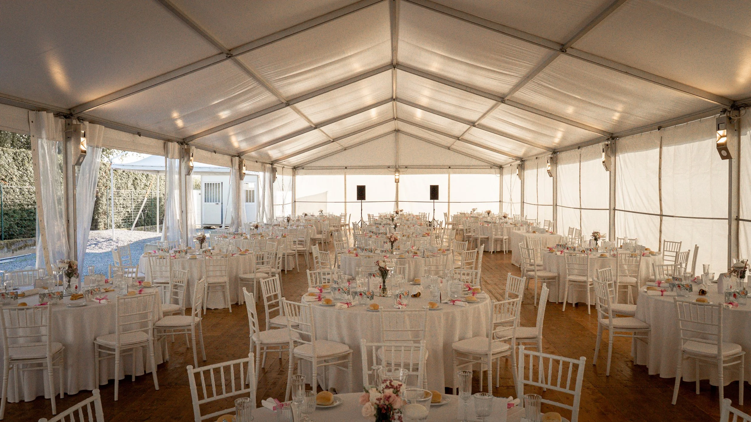 Interno di una tenda decorata per un evento, con tavoli tondi imbanditi con tovaglie bianche, piatti, bicchieri, posate e centrotavola floreali. Sedie biane disposte attorno ai tavoli.