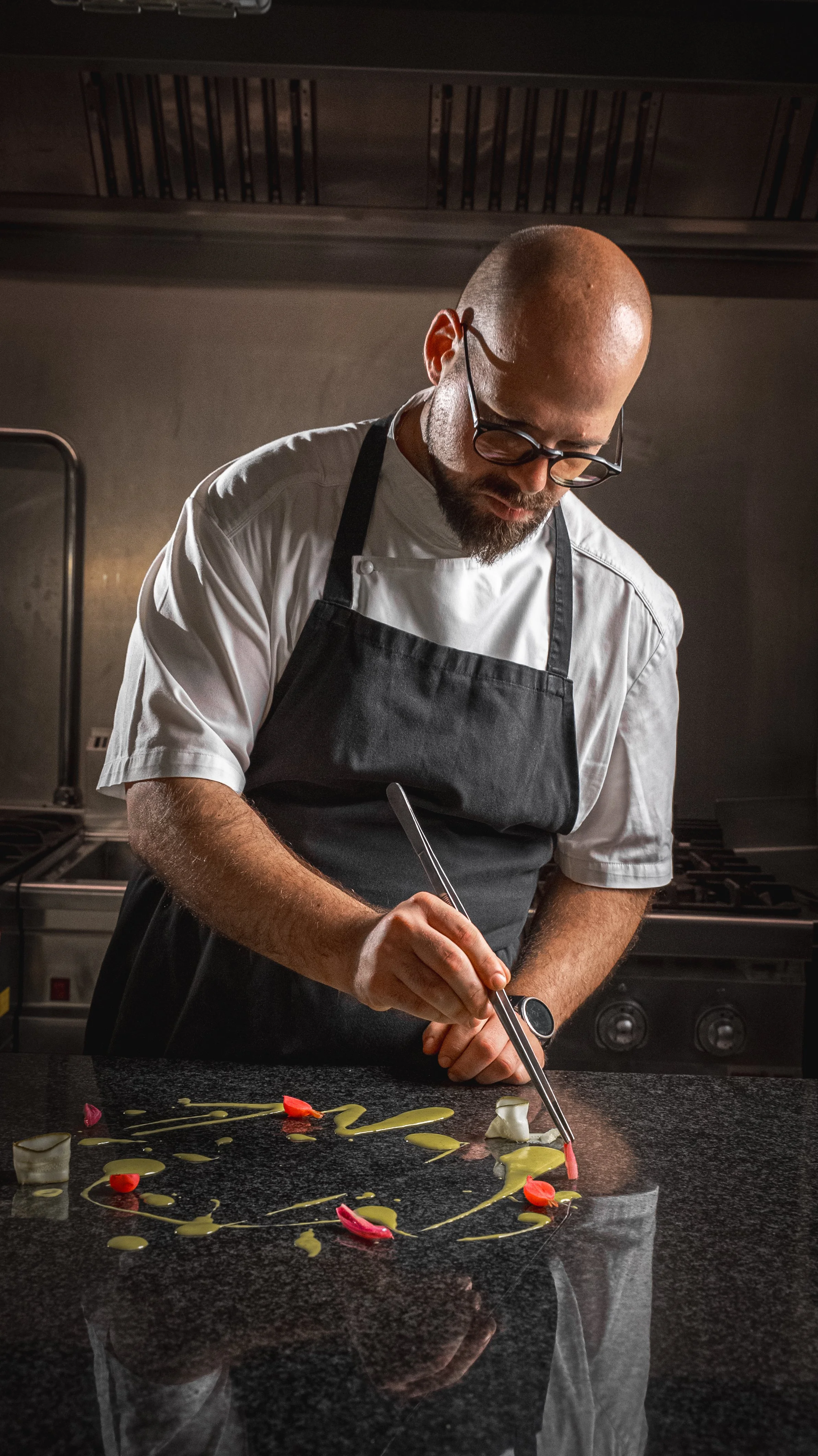 Chef professionista che prepara un piatto elegante in una cucina moderna, indossa un grembiule nero e occhiali, decorazioni in stile artistico sul piatto.
