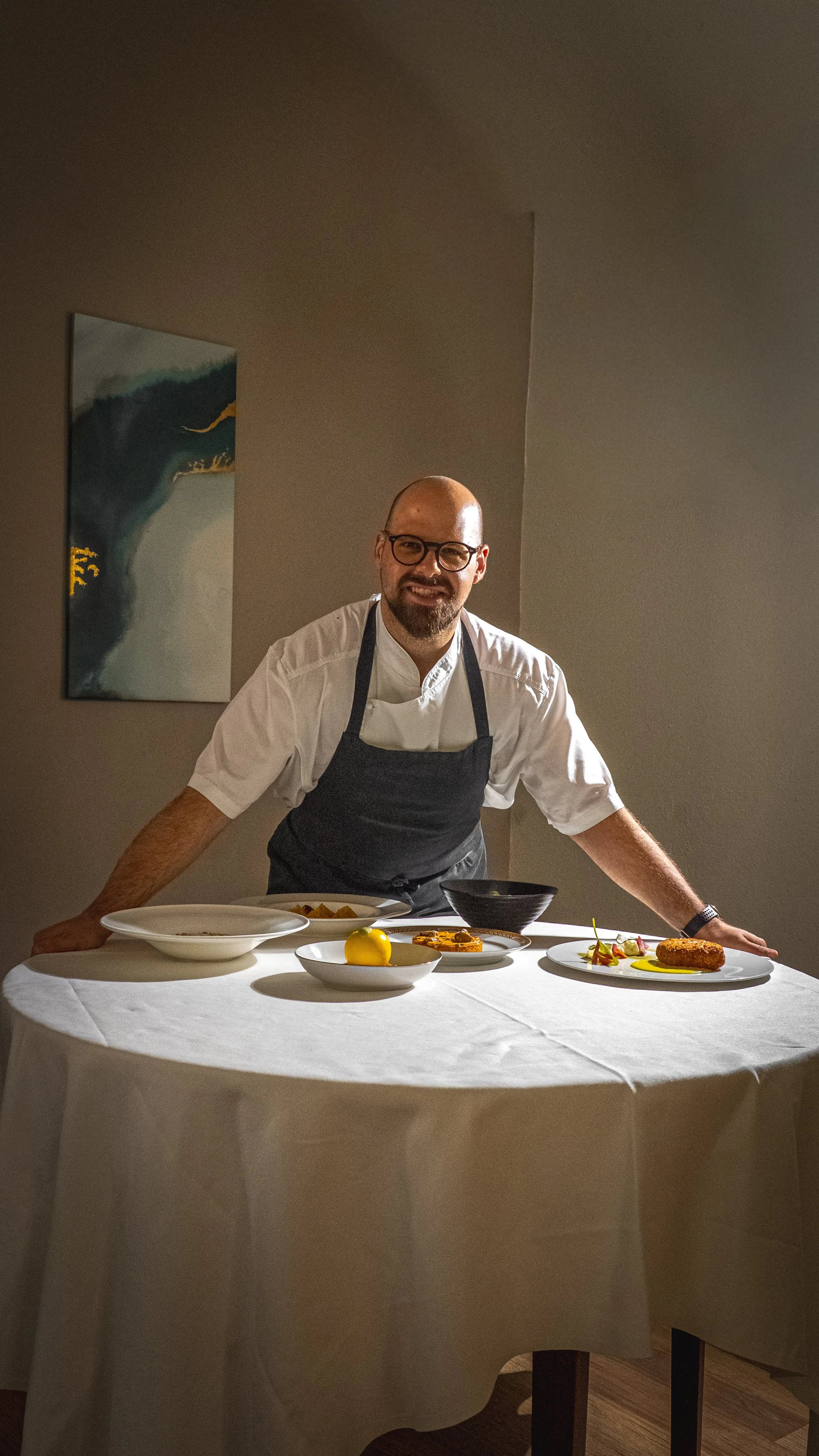 Chef sorridente davanti a piatti di alta cucina su tavolo tondo con tovaglia bianca.