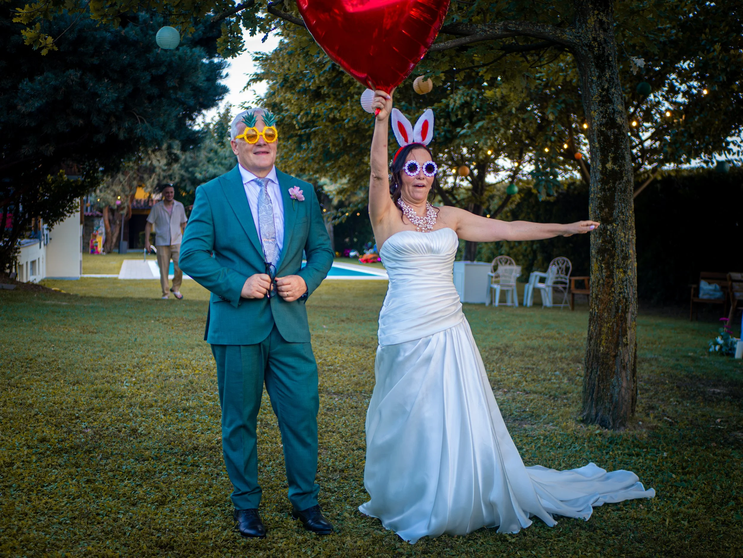 Due persone festeggiano all'aperto, una donna indossa un vestito bianco e occhiali con orecchie da coniglio, tiene un cuore rosso sfera, l'uomo indossa un completo blu, occhiali a forma di ananas e una camicia bianca.