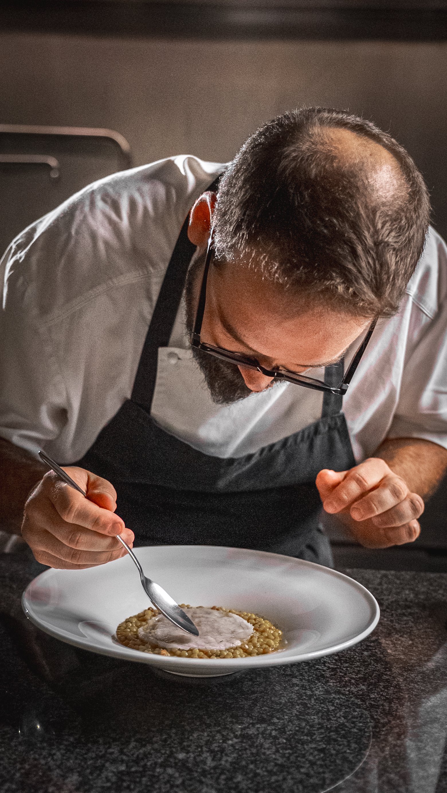 Un uomo con occhiali da vista, camice bianco e grembiule nero, sta decorando un piatto di cucina gourmet, aggrottato e concentrato mentre utilizza uno strumento per lavorare il cibo.