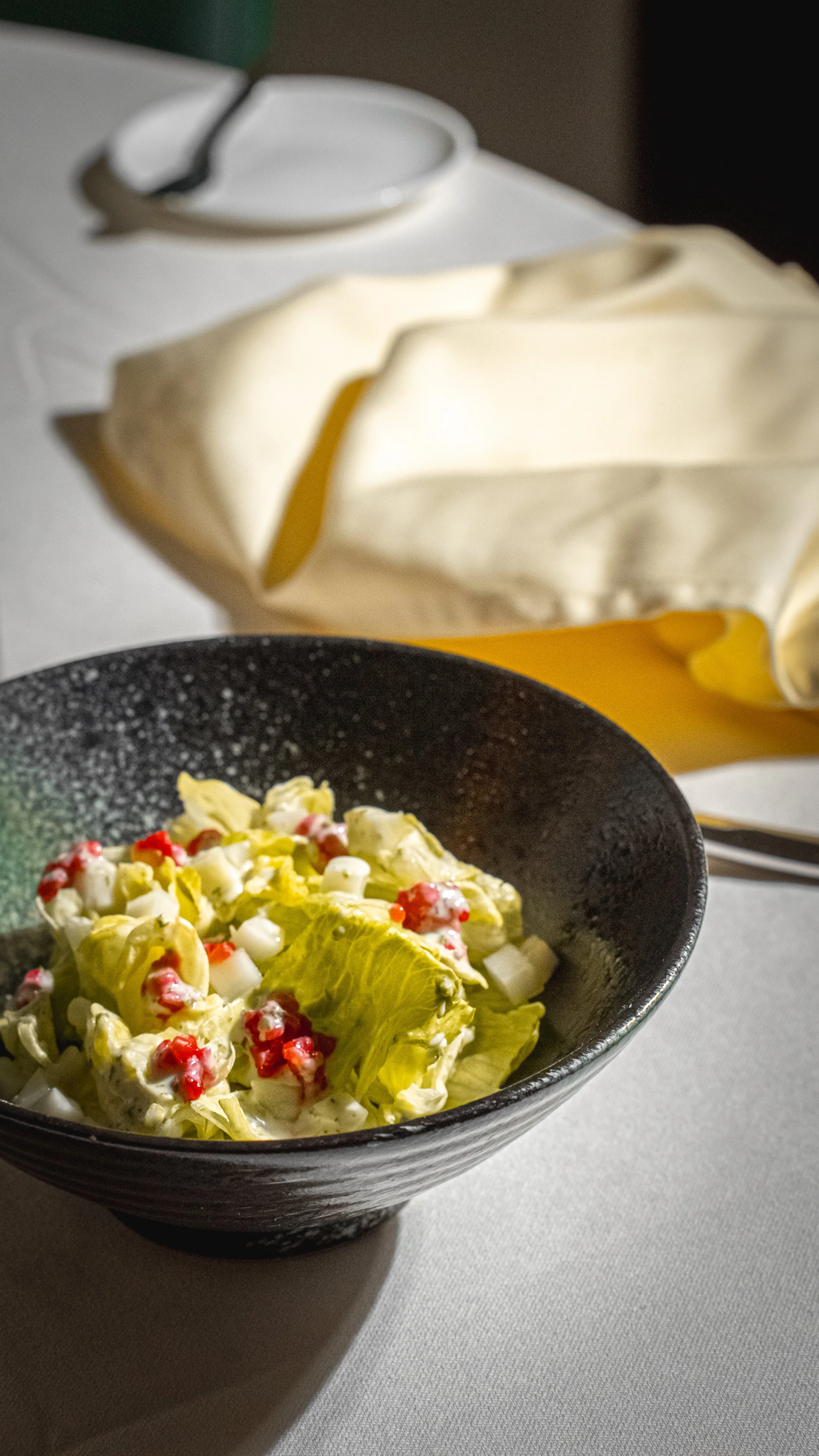 Insalata mista con lattuga, mais e semi di melograno in una ciotola nera, una forchetta e un coltello sul tavolo, uno sfondo scuro e una tovaglia bianca.