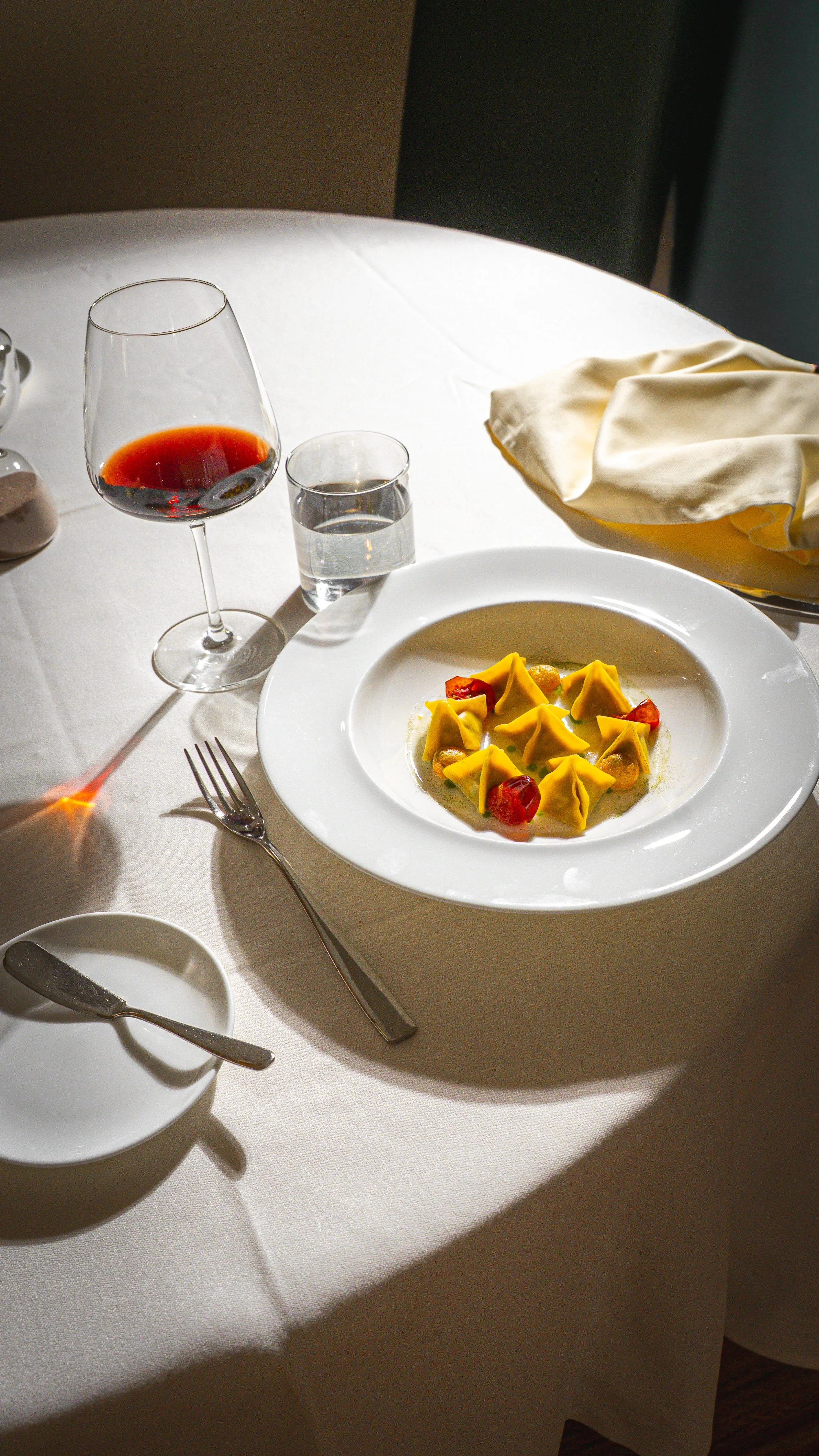 Un piatto di tortellini con pomodori ciliegini, un bicchiere di vino rosso, un bicchiere di acqua e una tovaglia bianca su un tavolo apparecchiato in un ristorante.