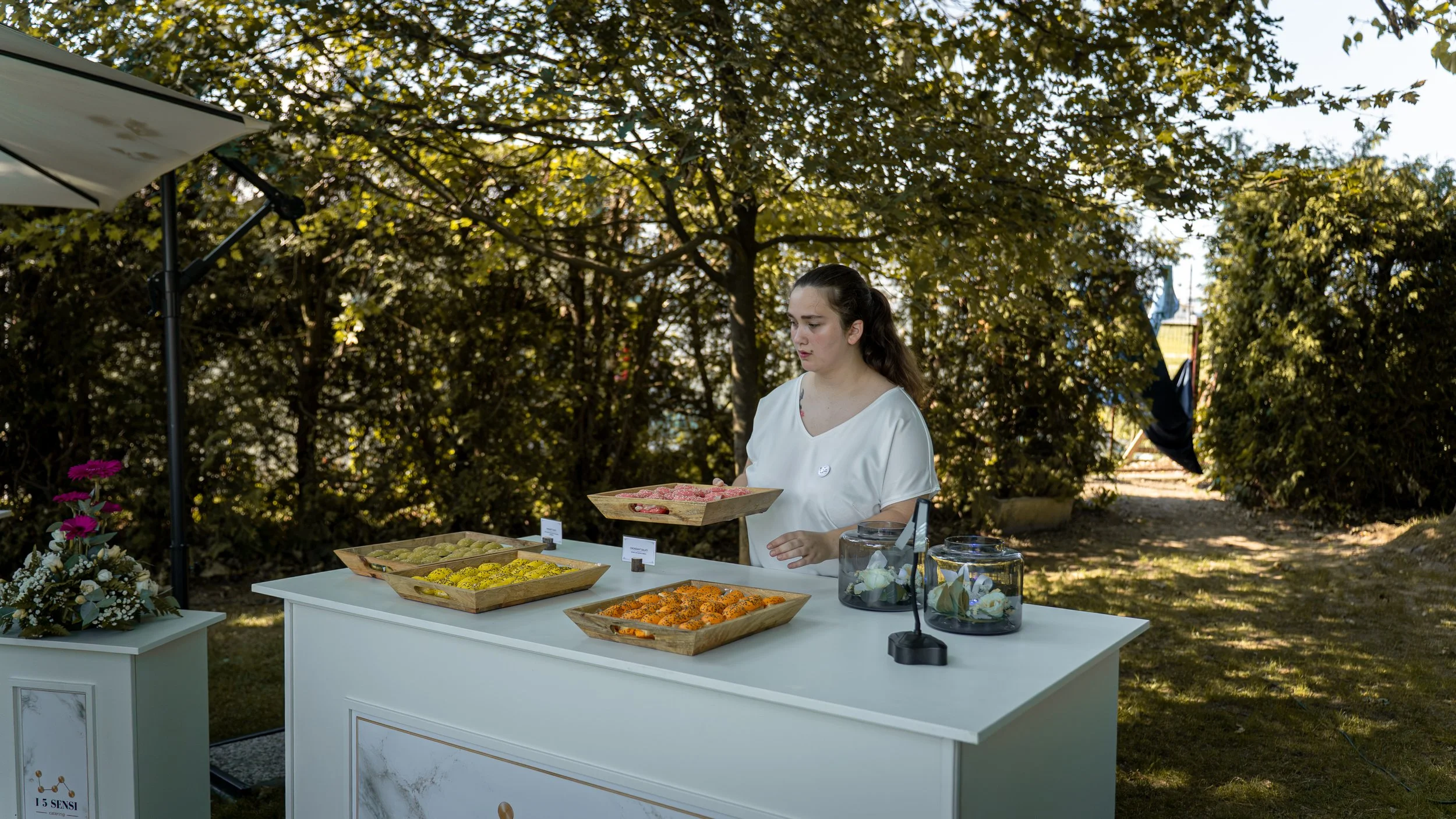 Una giovane donna che vende dolci colorati su un banco all'aperto circondata da alberi con luce solare.