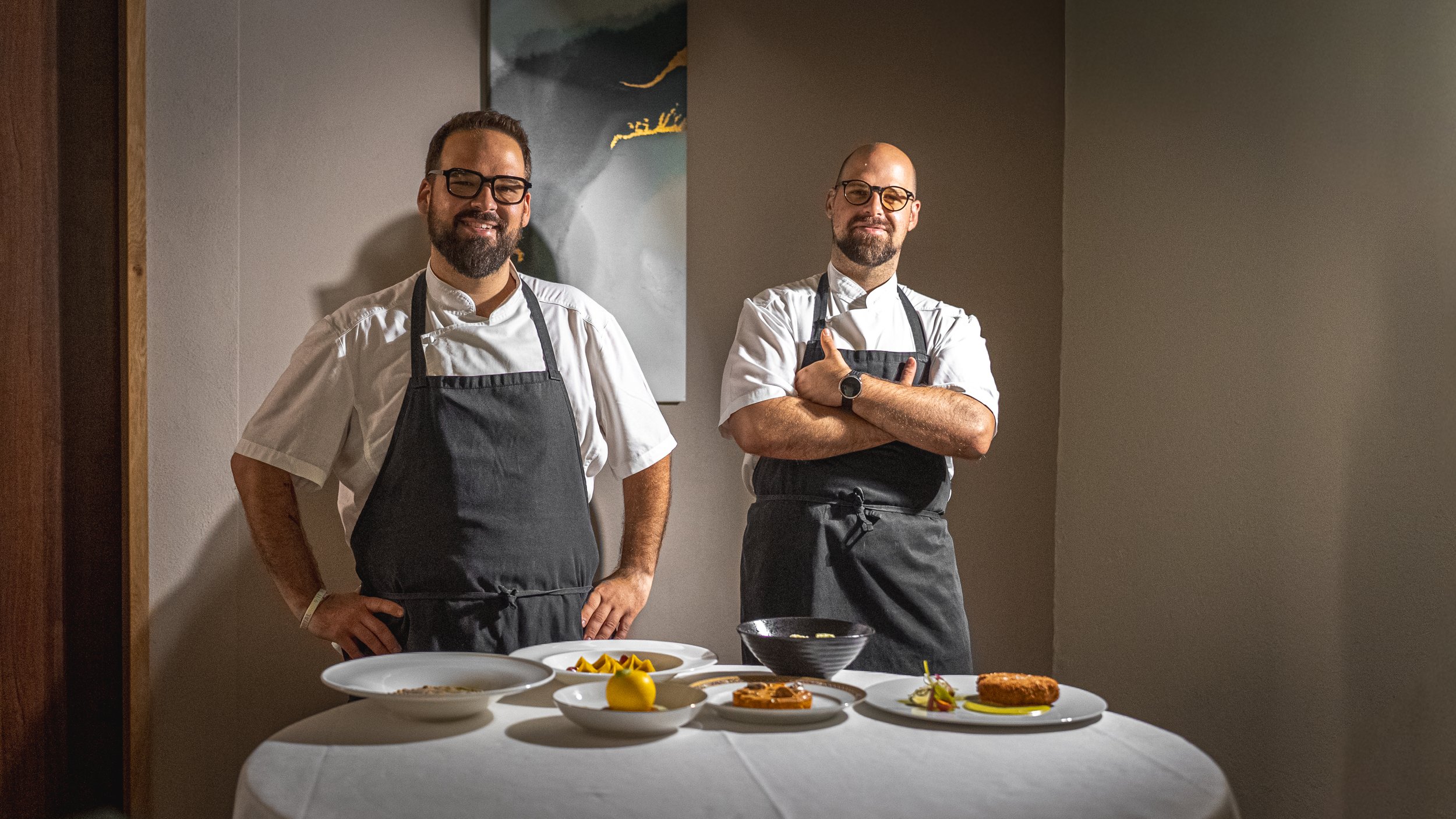 Due uomini chef in divisa, uno con barba e altri senza, sorridenti, indossano grembiuli neri e sono in una cucina o sala da cena. In primo piano, un tavolo con piatti di cibo elaborato, tra cui dessert e piatti decorati.