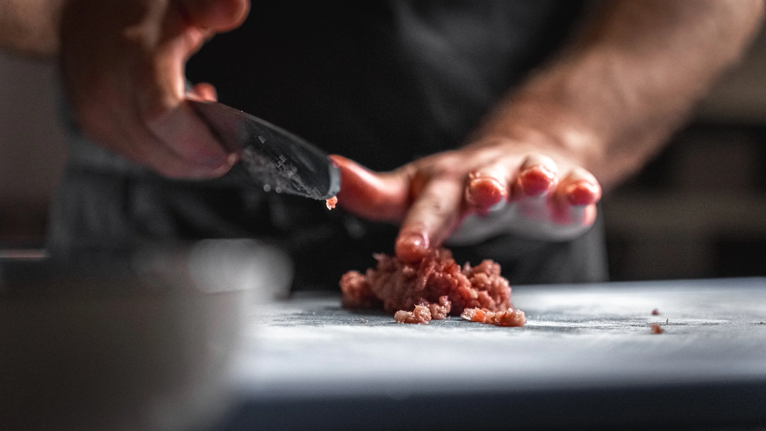 Una mano che taglia carne macinata con un coltello, con altra mano che tiene la carne. Sul tavolo c'è carne macinata e un coltello.
