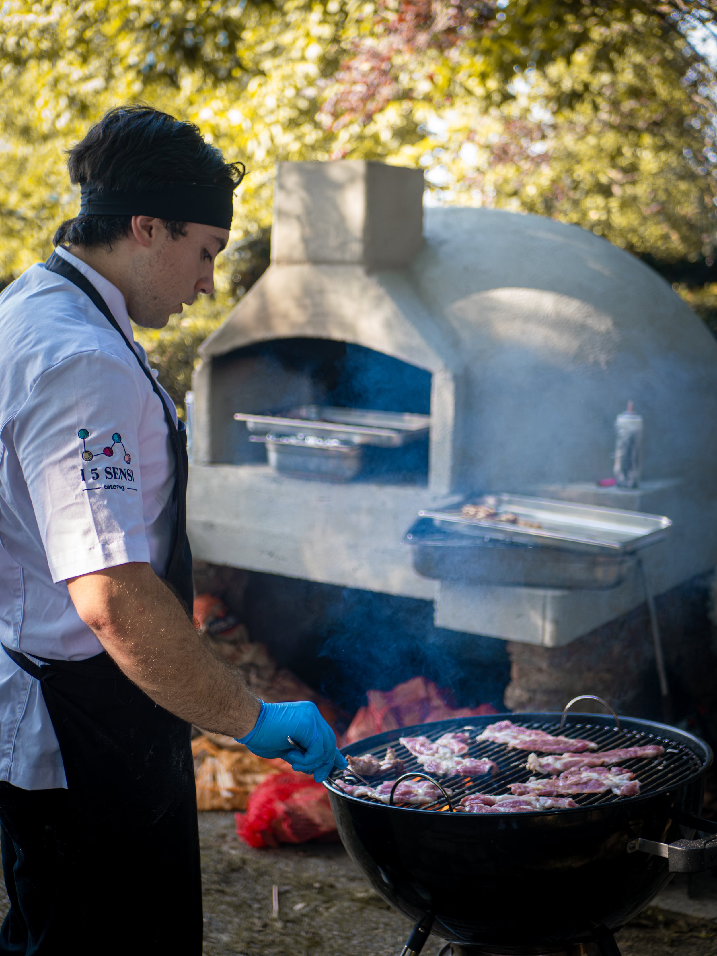 Un giovane cuoco mentre arrostisce carne sul barbecue all'aperto, con un grande forno di pietra sullo sfondo e alberi con foglie autunnali.