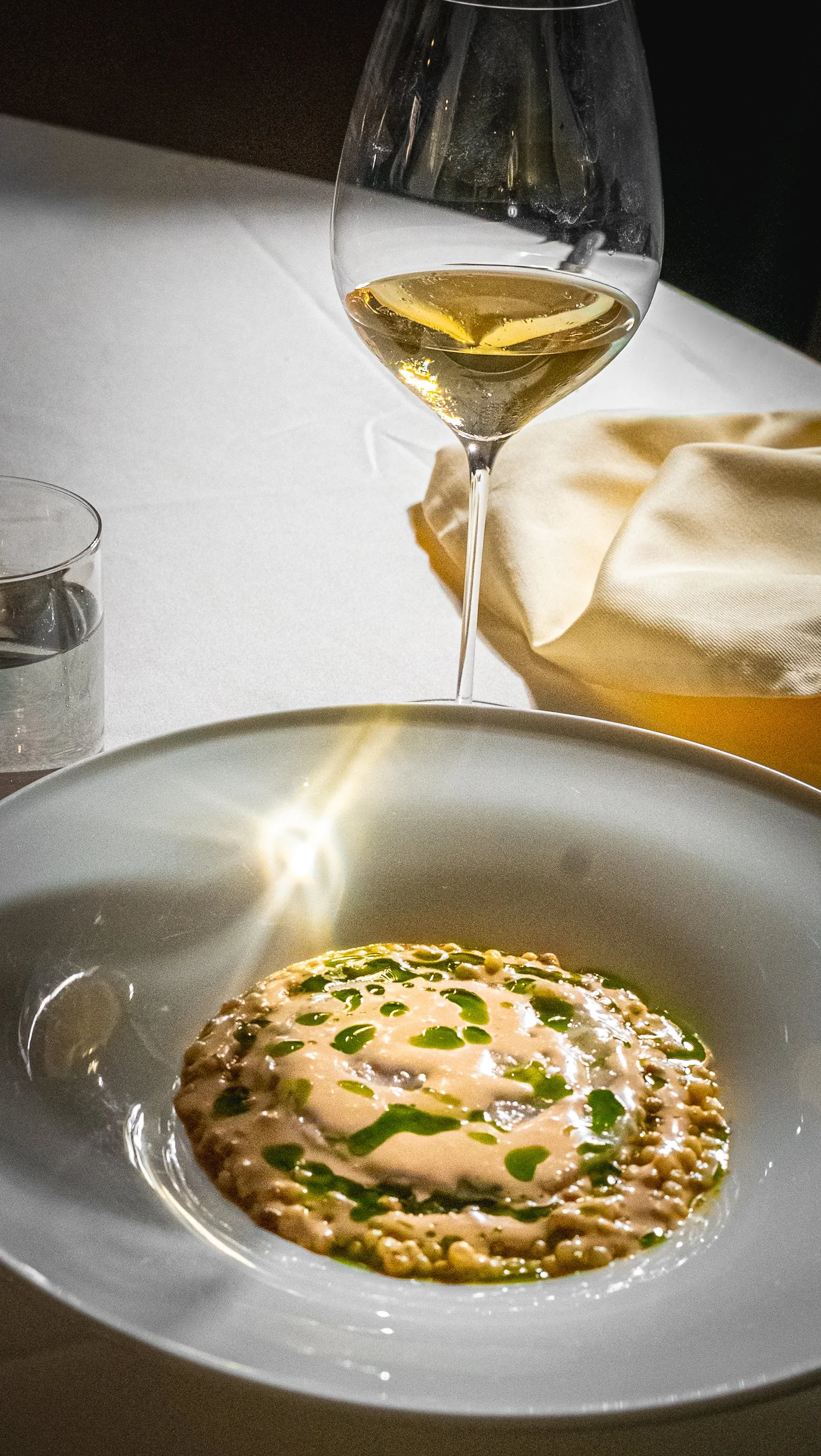 Un bicchiere di vino bianco sul tavolo, con una fetta di limone all'interno, e un piatto con un risotto decorato con olio e prezzemolo.