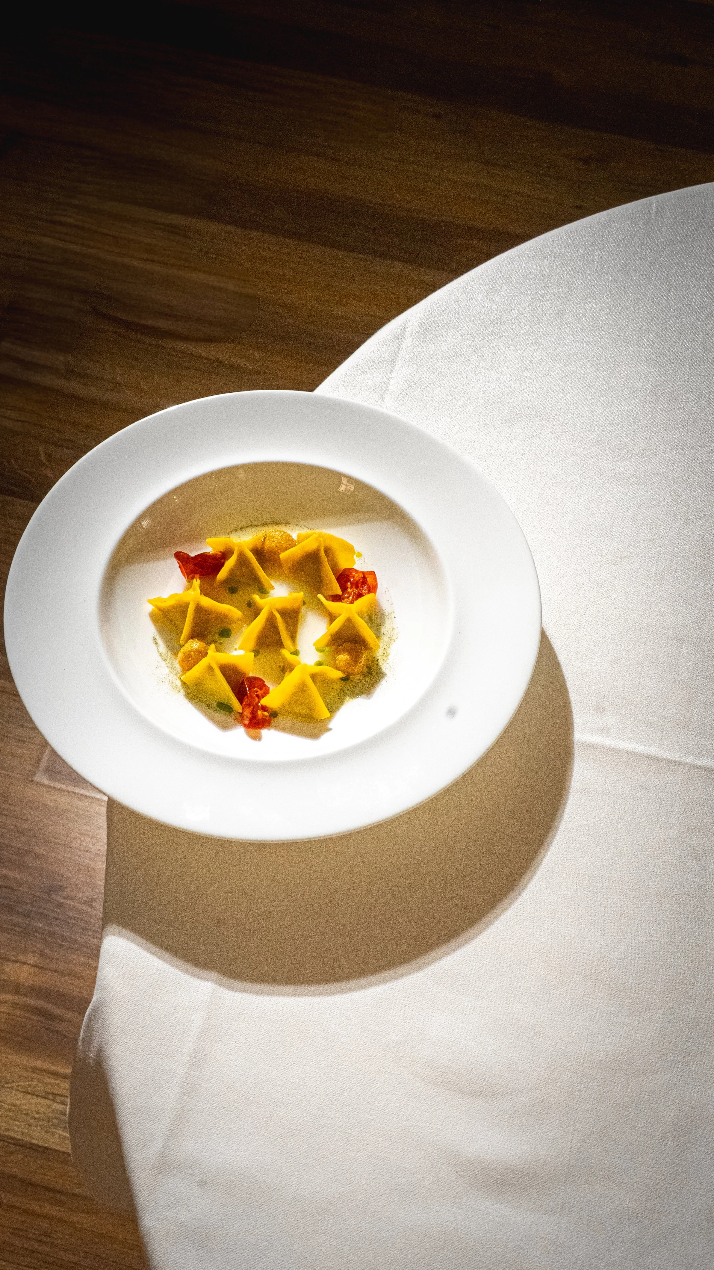Piatto raffinato con ravioli gialli accompagnati da pomodori secchi, conditi con salsa e decorazioni verdi, presentato in una ciotola bianca.