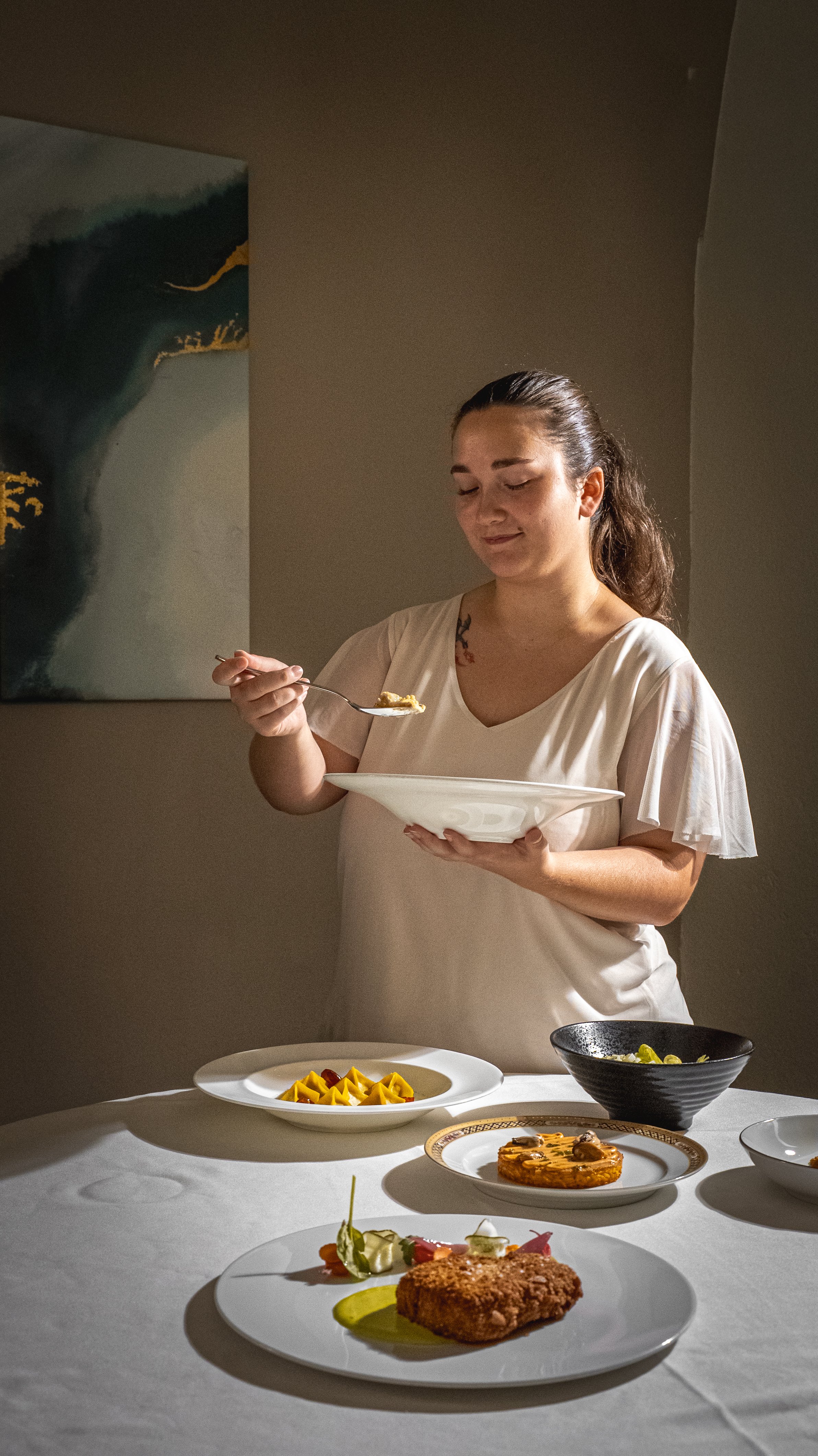 Una donna con capelli raccolti e vestito bianco che si sta preparando a mangiare, con piatti di cibo su un tavolo apparecchiato.
