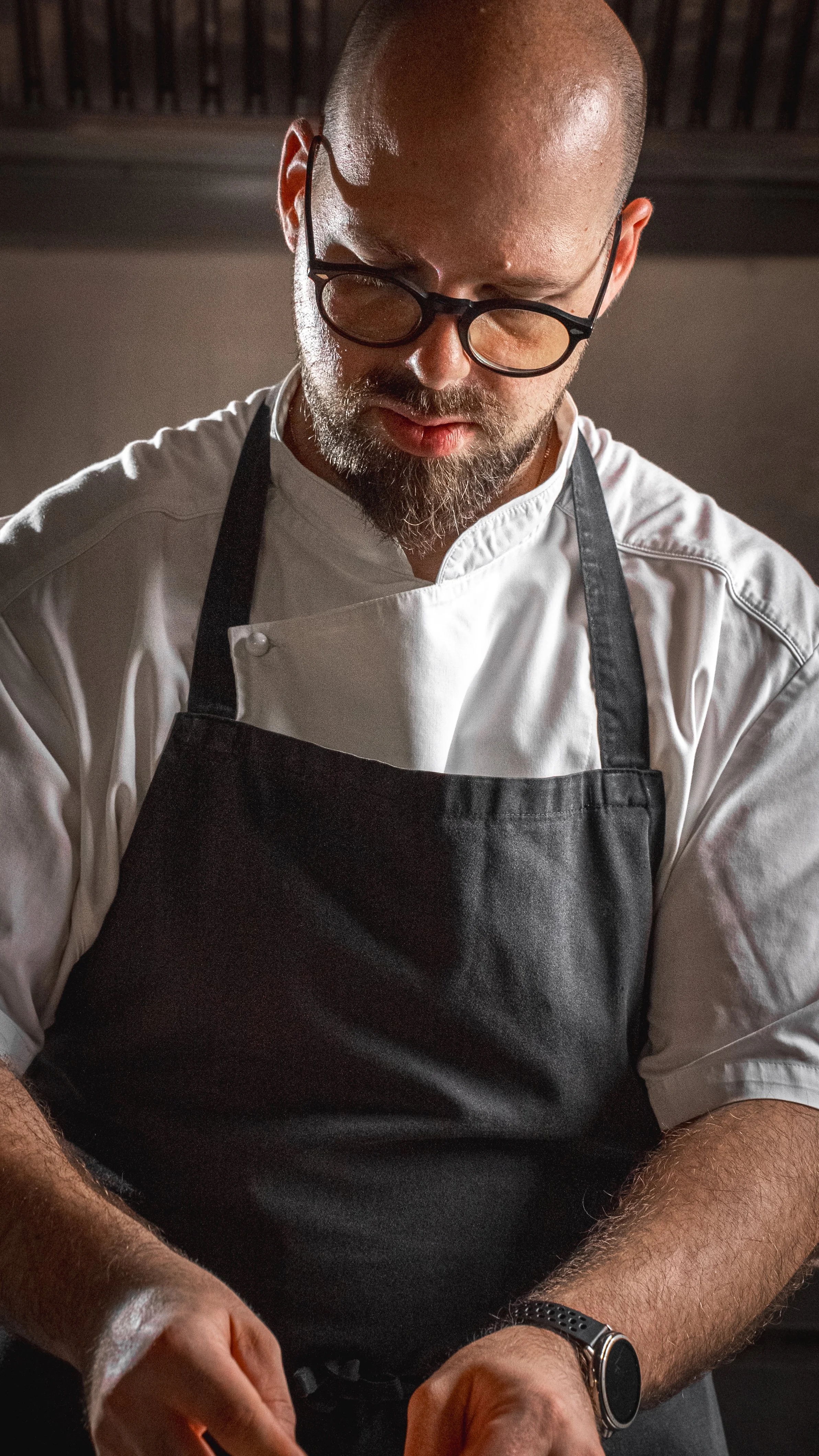Un uomo con occhiali, barba e cappello che indossa una giacca bianca e un grembiule nero, intento a cucinare.