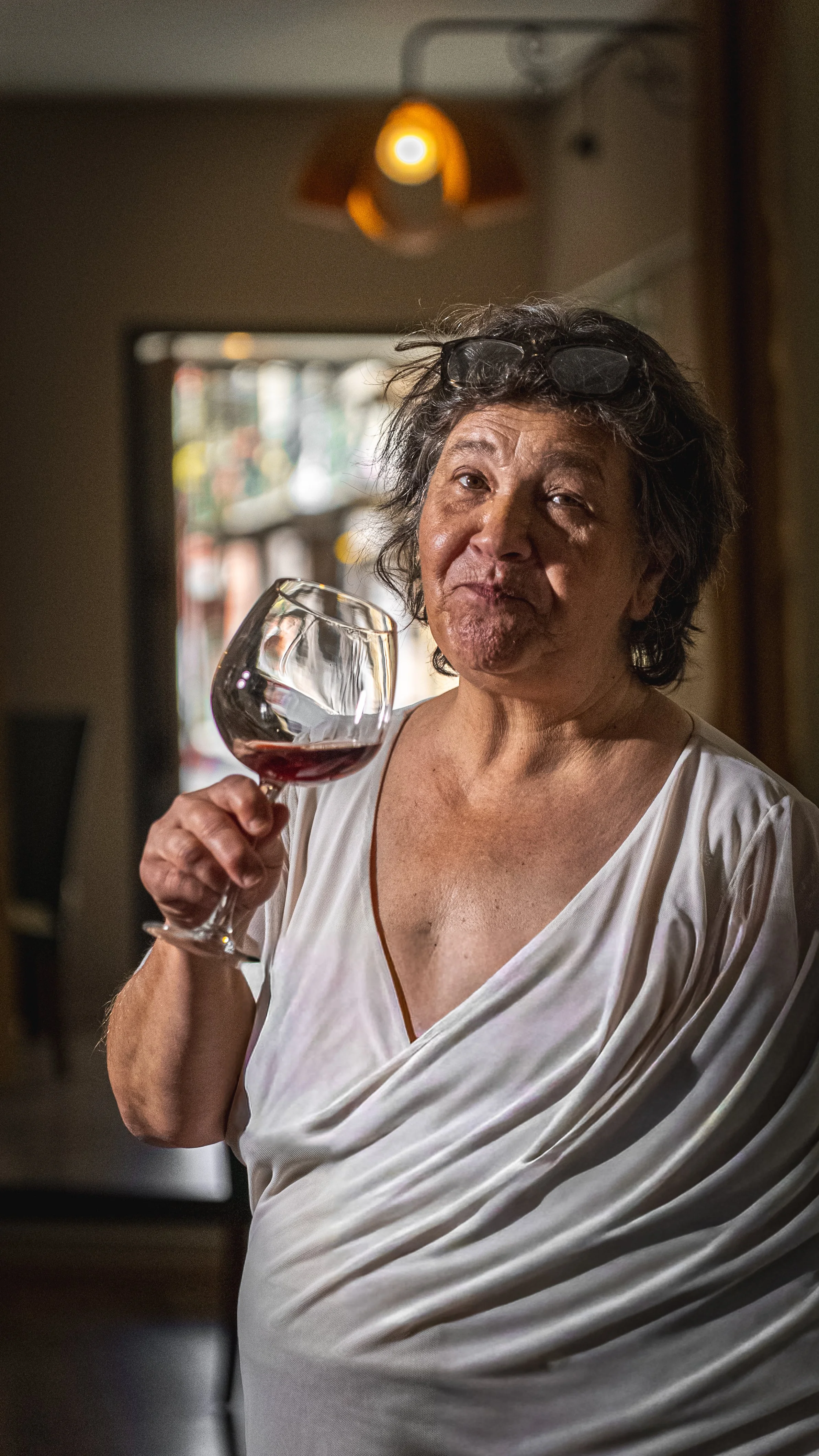 Donna anziana con occhiali sulla testa che tiene un bicchiere di vino rosso, vestita con una camicia bianca in un ambiente domestico.