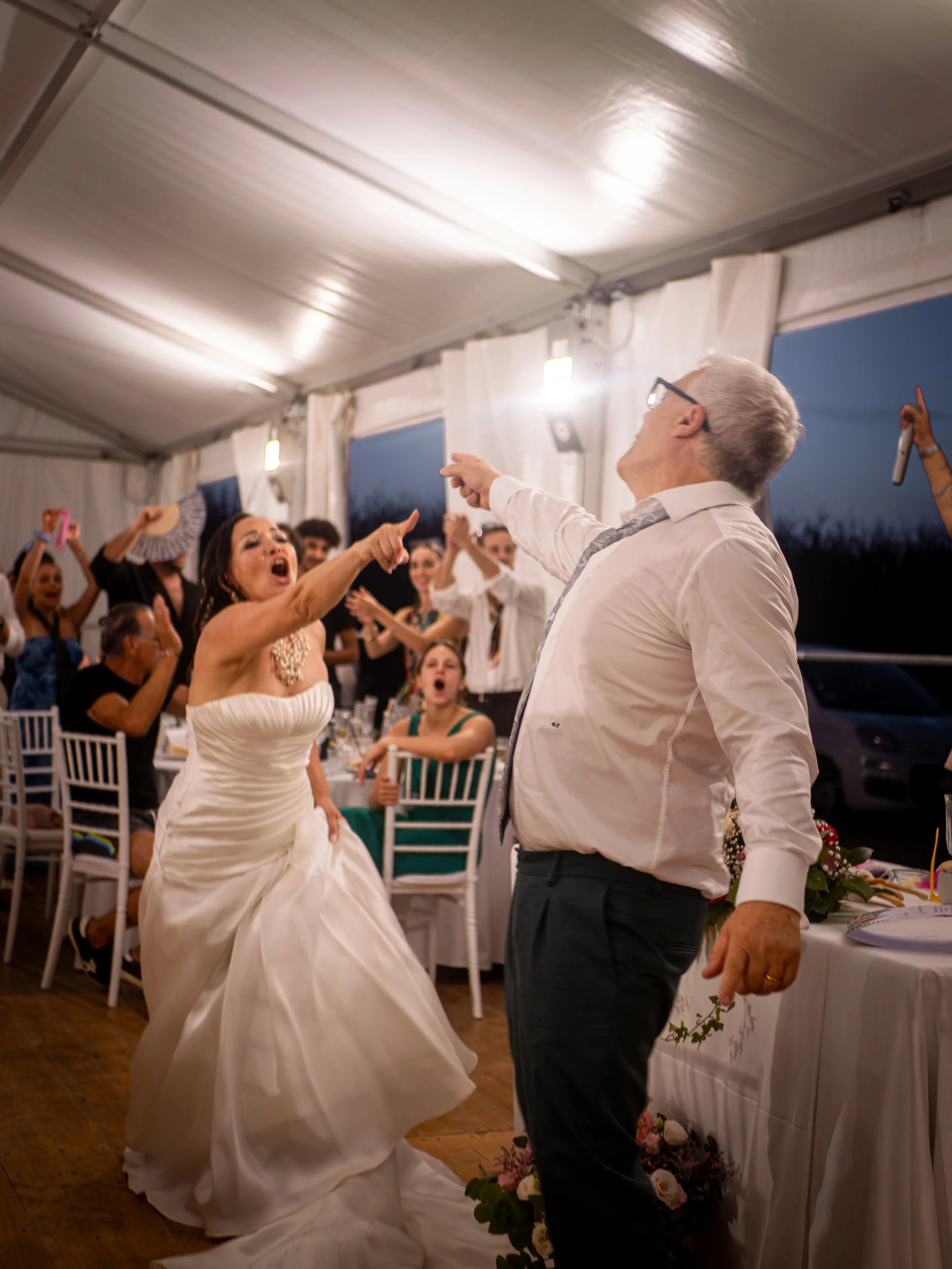 Una coppia di sposi balla durante il matrimonio, circondati da amici e parenti in un ambiente festoso. La sposa indossa un abito bianco e il marito una camicia bianca con cravatta.