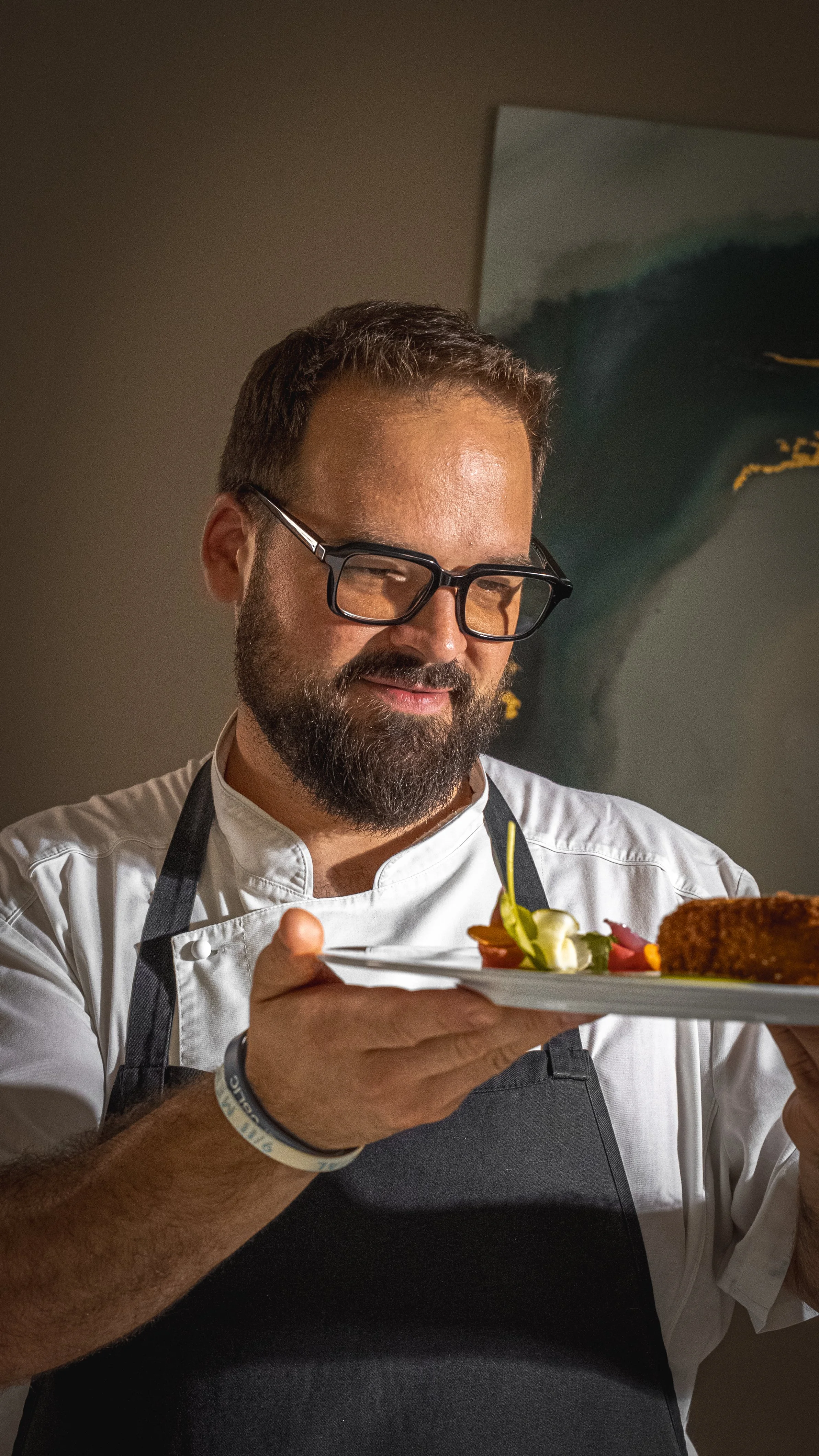 Un uomo con occhiali e barba, vestito con una giacca bianca, tiene un piatto di cibo decorato, in un ambiente interno.