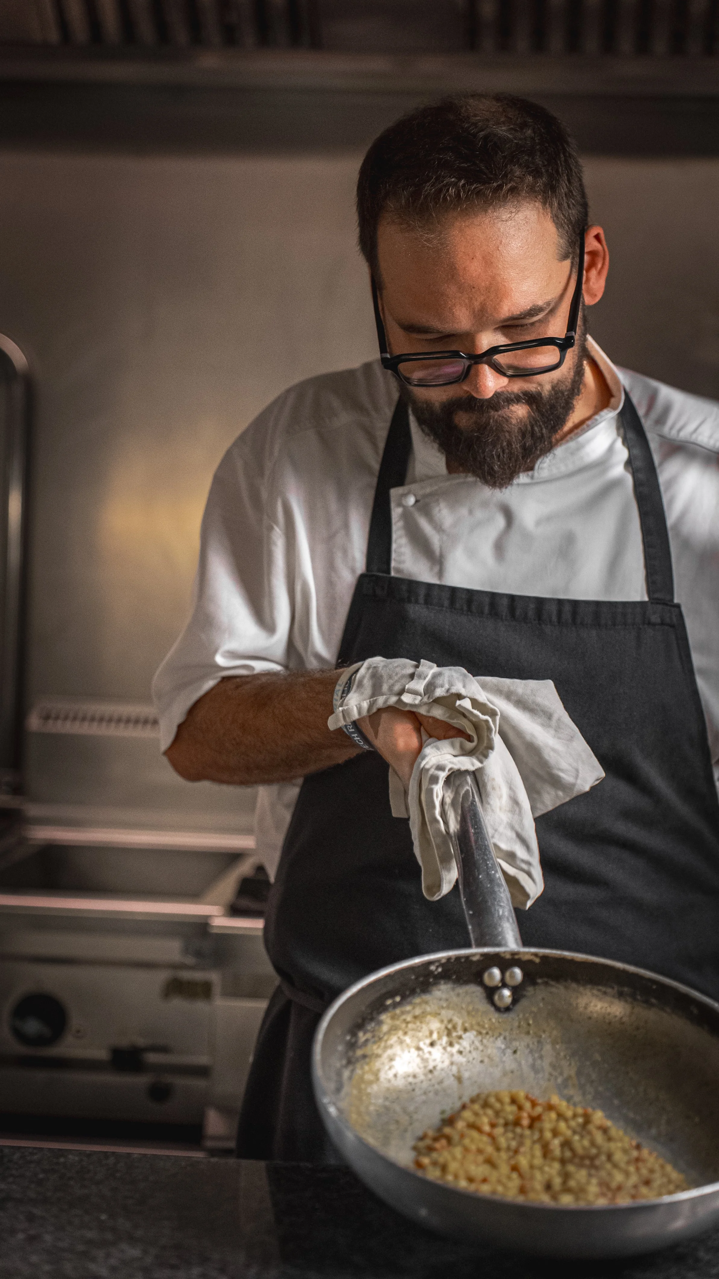 Un uomo con occhiali da vista, barba e pettinatura cortissima, indossa una giacca bianca e un grembiule nero, e sta cucinando in una cucina moderna. Tiene una spatola e un grembiule appoggiato attorno al collo, mentre mescola ingredienti nel tegame.