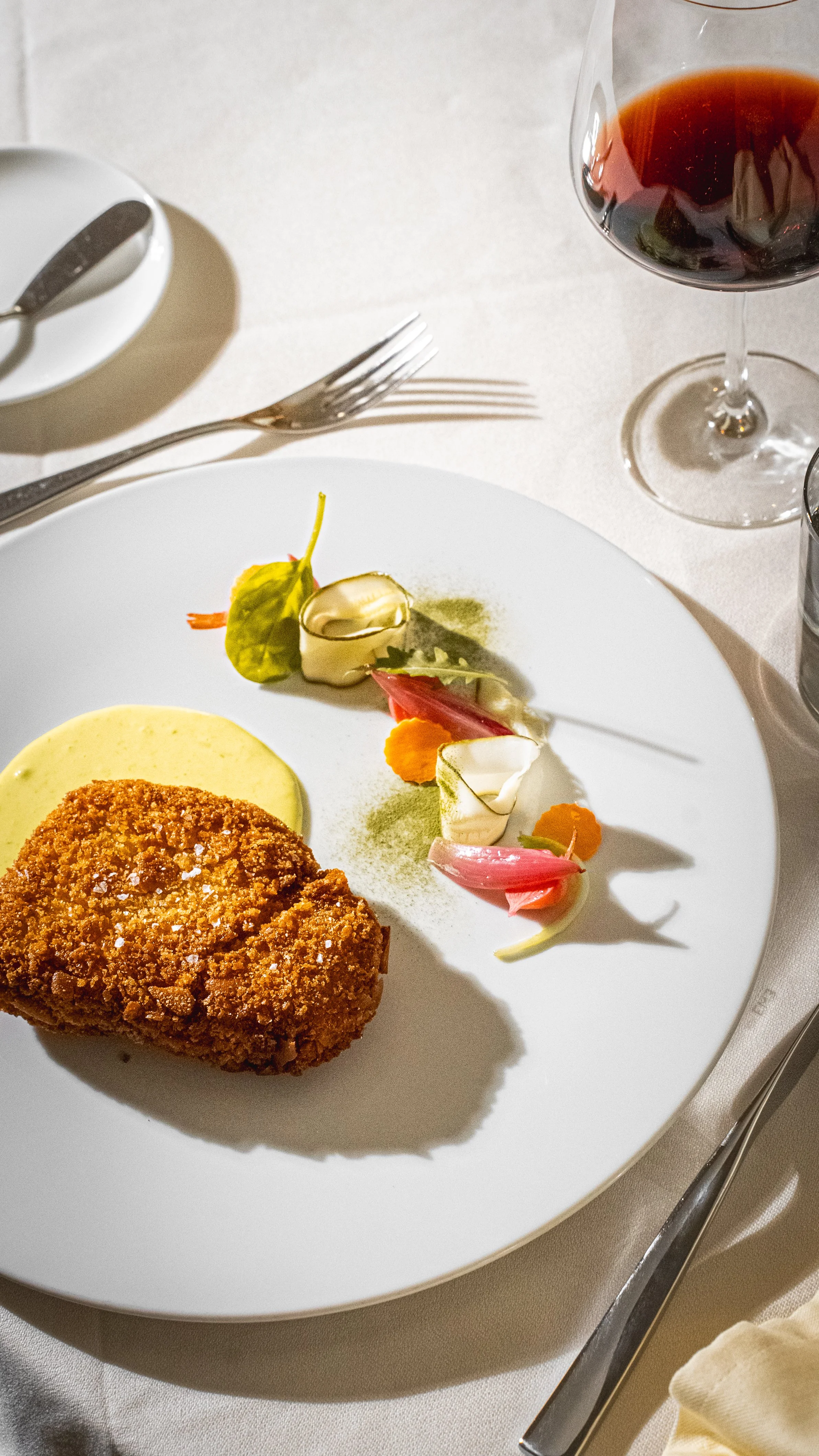 Piatti di cibo raffinato su un piatto bianco, con un pezzo di carne impanato, salse e decorazioni di verdure, accompagnato da un bicchiere di vino rosso.