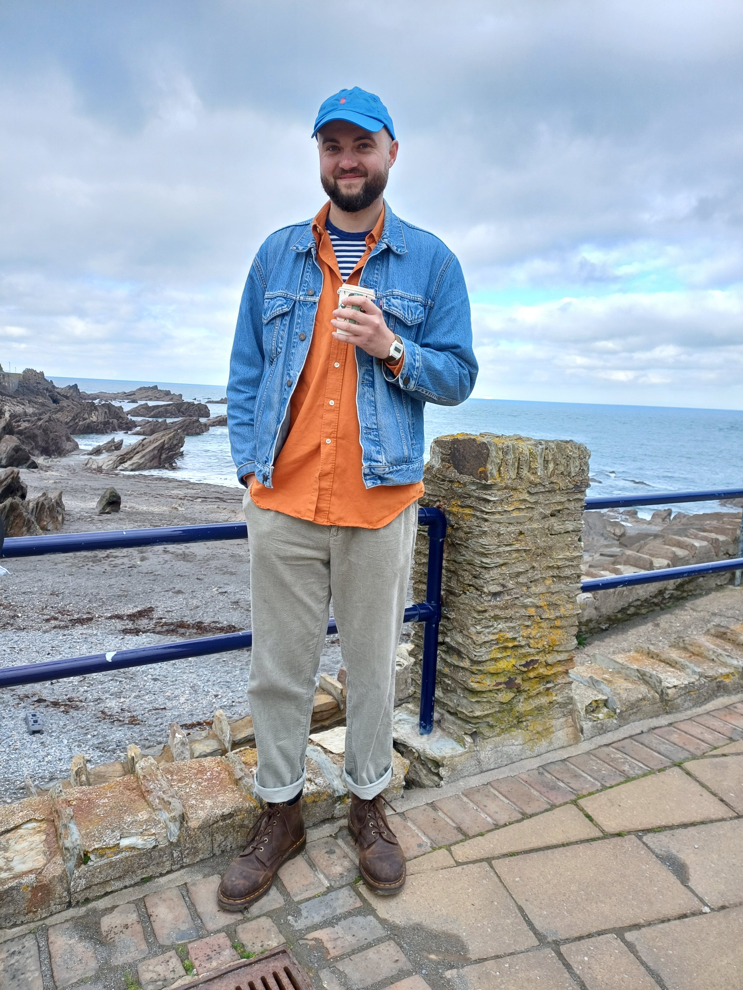 A man with a beard wearing a blue cap, denim jacket, and beige pants, standing by the ocean with rocks and a cloudy sky in the background, holding a coffee cup.