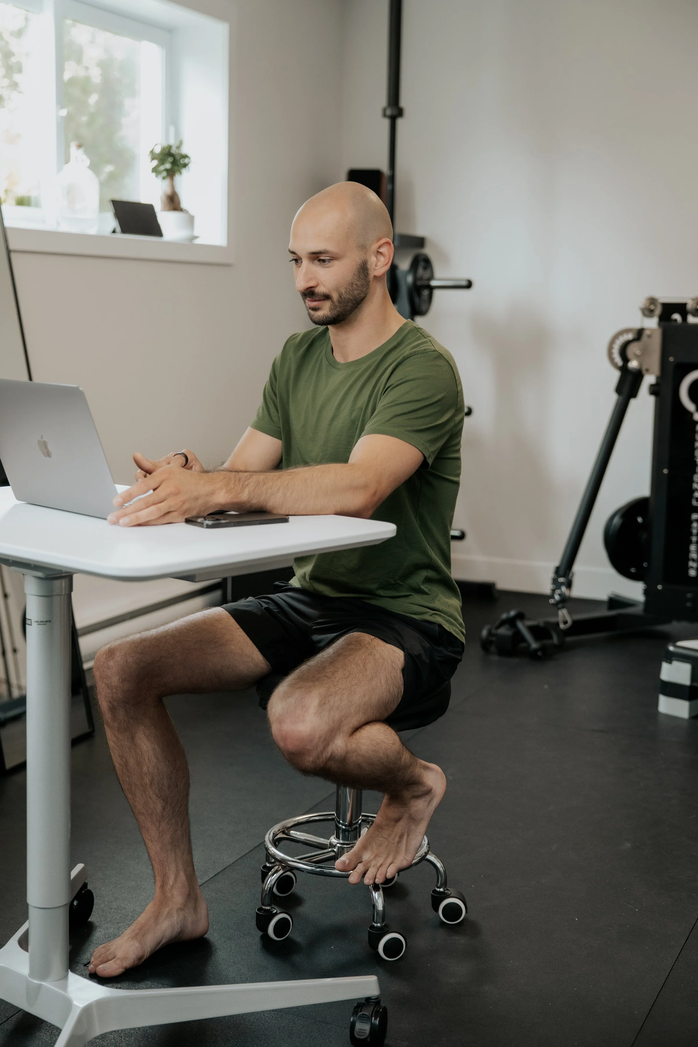 Un homme à tête chauve et barbe, habillé d'un t-shirt vert et d'un short noir, est assis sur une chaise derrière un bureau blanc, travaillant sur un ordinateur portable. Il est pieds nus, avec un pied posé sur la barre de la chaise et l'autre sur le sol. En arrière-plan, on voit des équipements de musculation dans une salle d'entraînement.