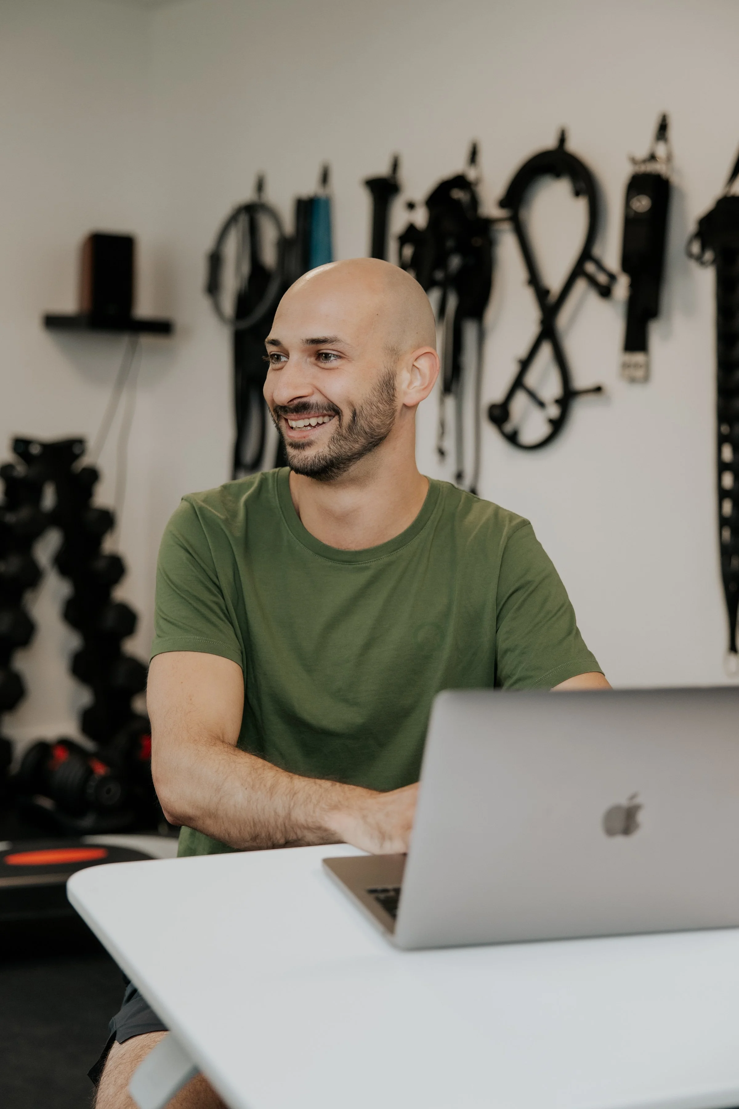 Homme barbu souriant assis devant un ordinateur portable dans une salle de sport ou un centre de fitness avec des équipements de musculation en arrière-plan.