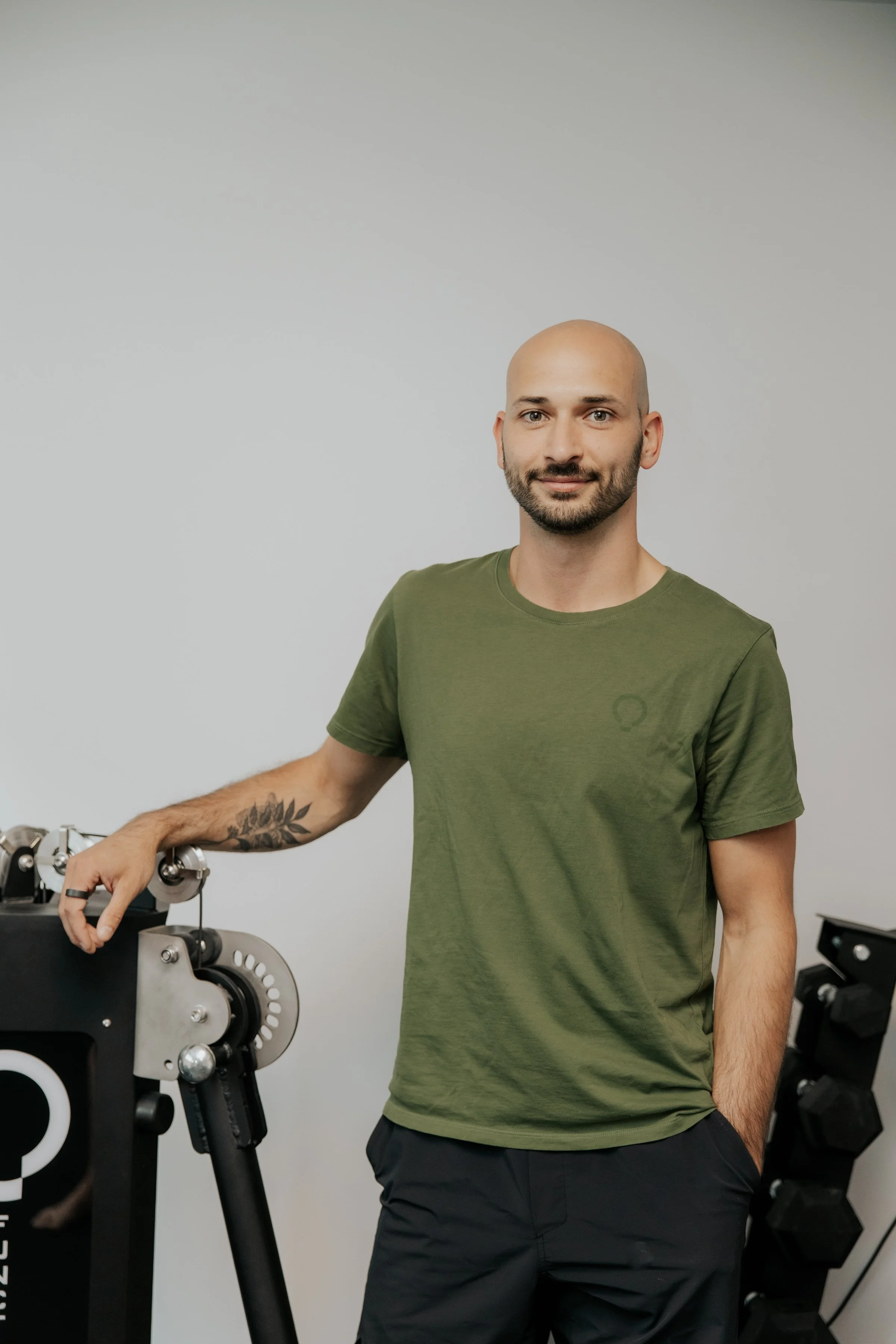 Un homme chauve avec une barbe souriante portant un t-shirt vert, debout à côté d'une machine, dans une pièce avec un mur blanc.