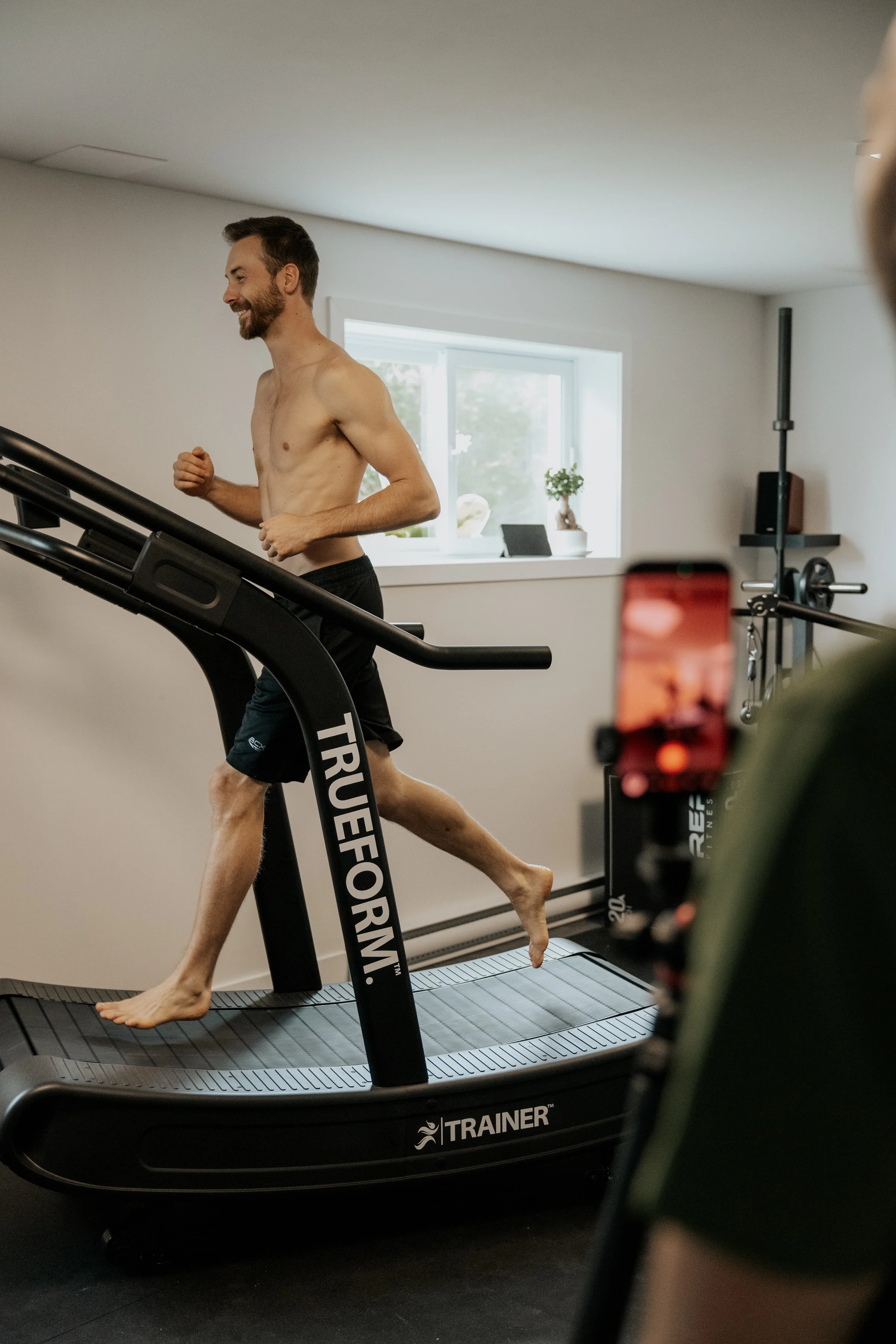 Un homme en short court court sur un tapis roulant de fitness, il est torse nu, souriant, dans une pièce bien éclairée avec une fenêtre. Une personne filme avec son téléphone.