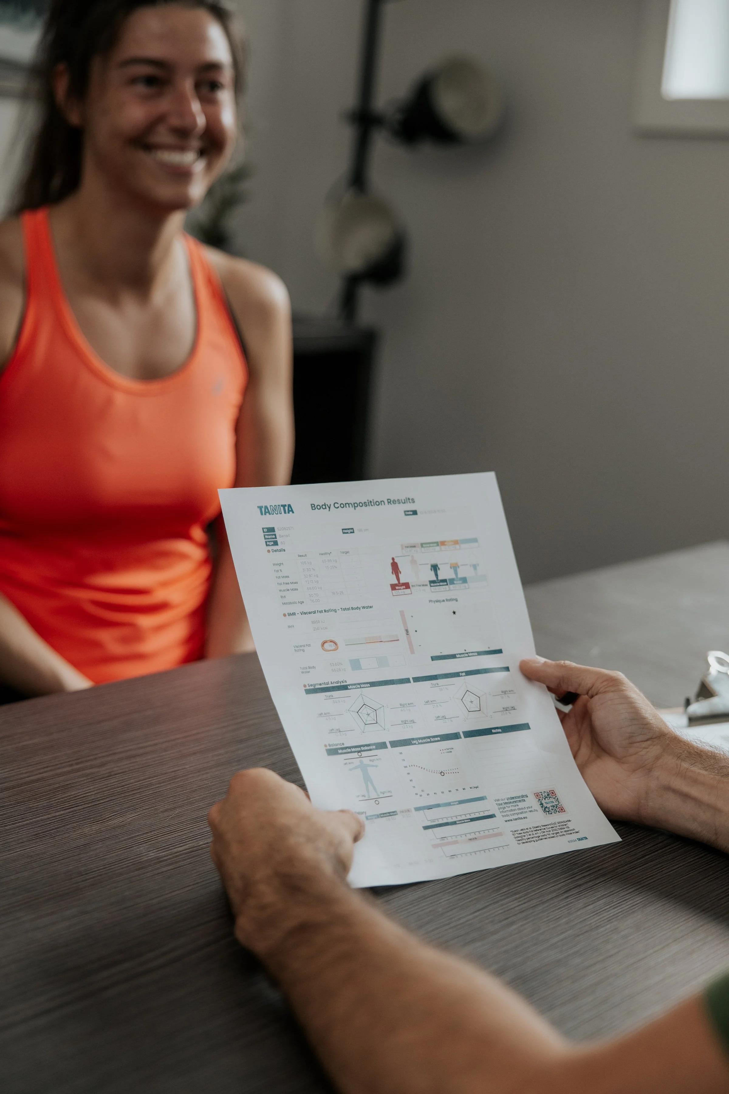 Une femme souriante en haut à gauche, portant un t-shirt orange, devant une fenêtre. Elle reçoit un rapport contenant des résultats d'évaluation corporelle, démonstration graphique et statistiques.