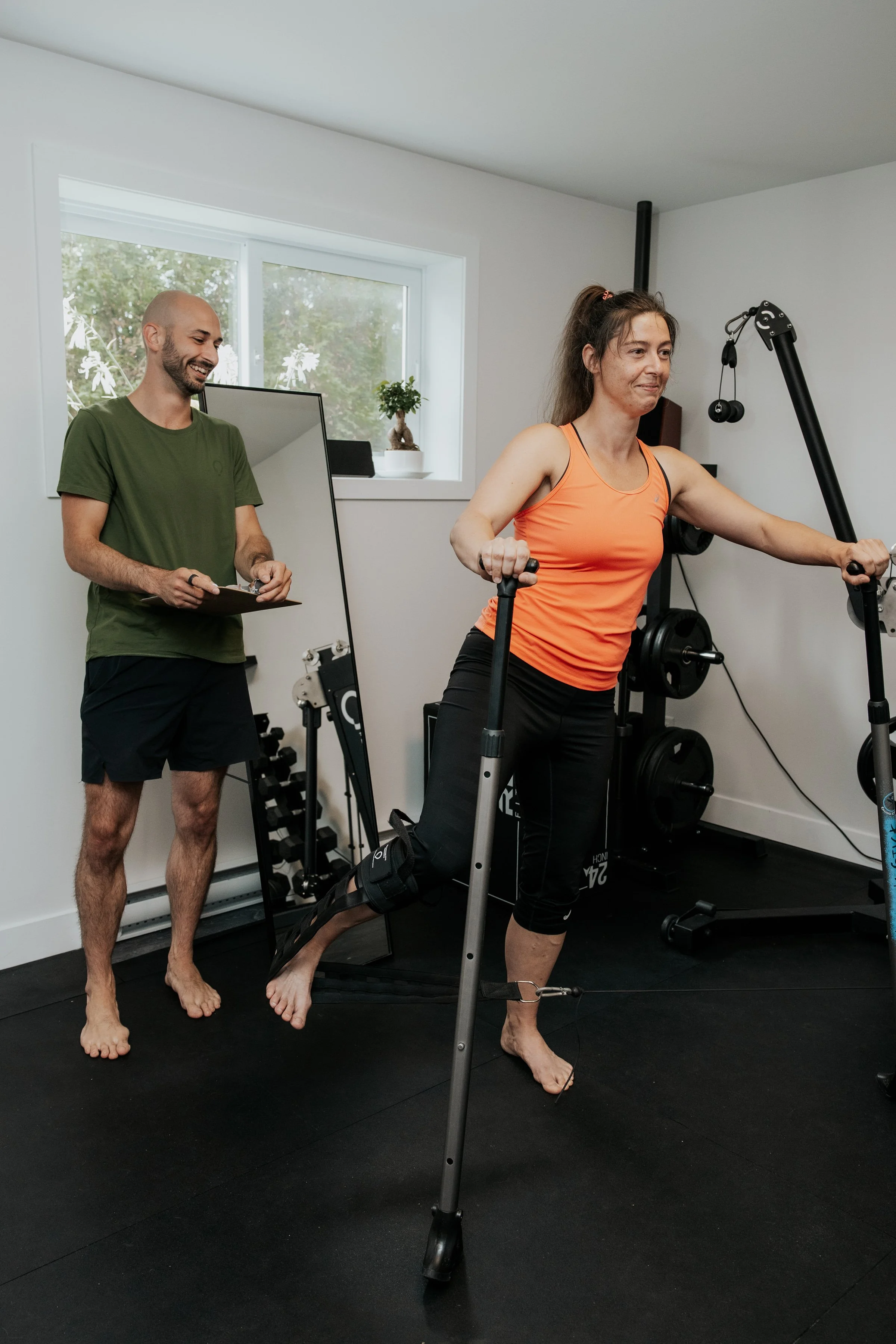 Une femme en tenue de sport utilise un rameur dans une salle d'entraînement, un homme observe et note quelque chose sur un clip-board. La salle est lumineuse avec une fenêtre et des décorations végétales.