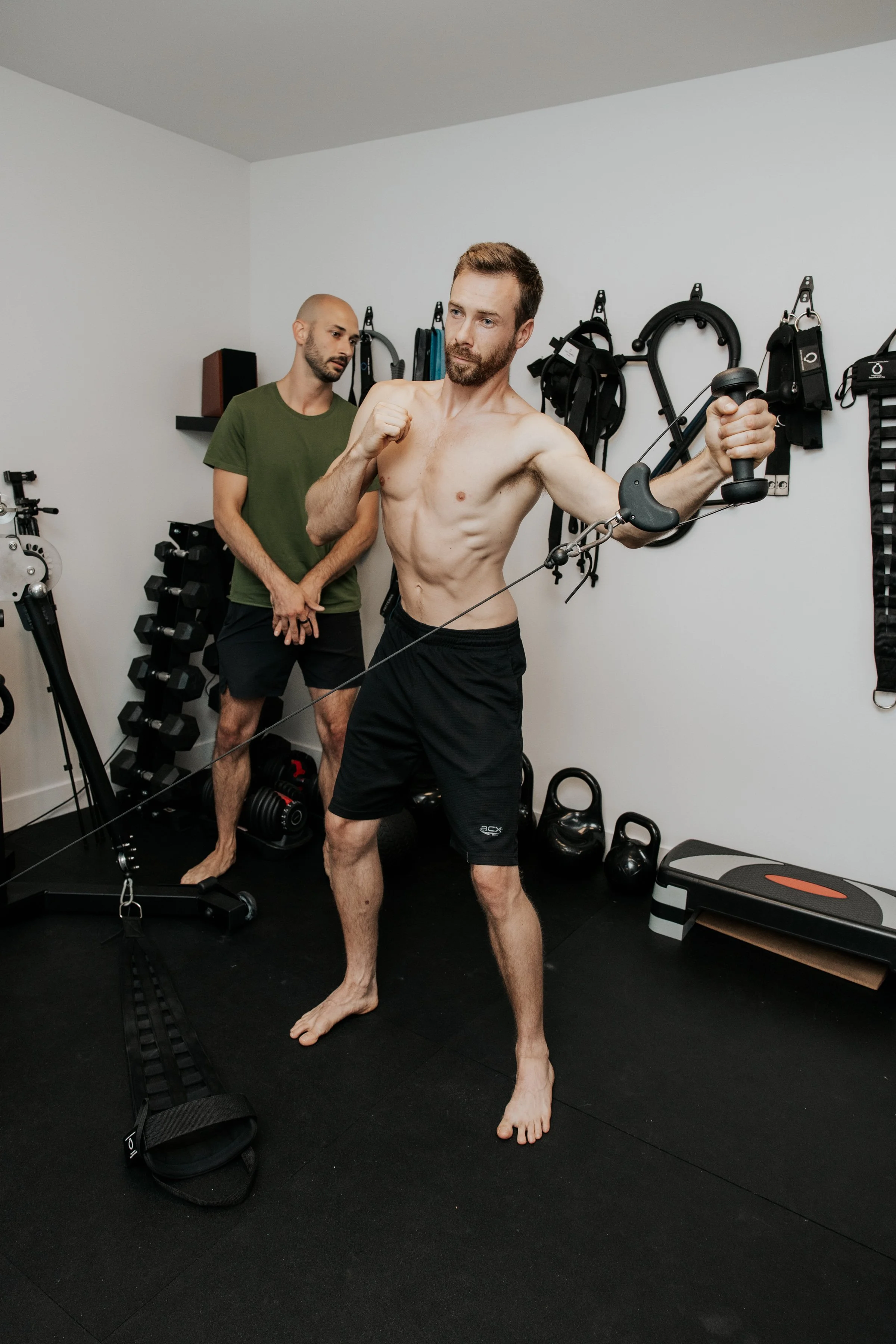 Un homme musclé posant avec un poing levé dans une salle d'entraînement, un autre homme le regarde. La salle a des équipements de musculation sur le mur et au sol.