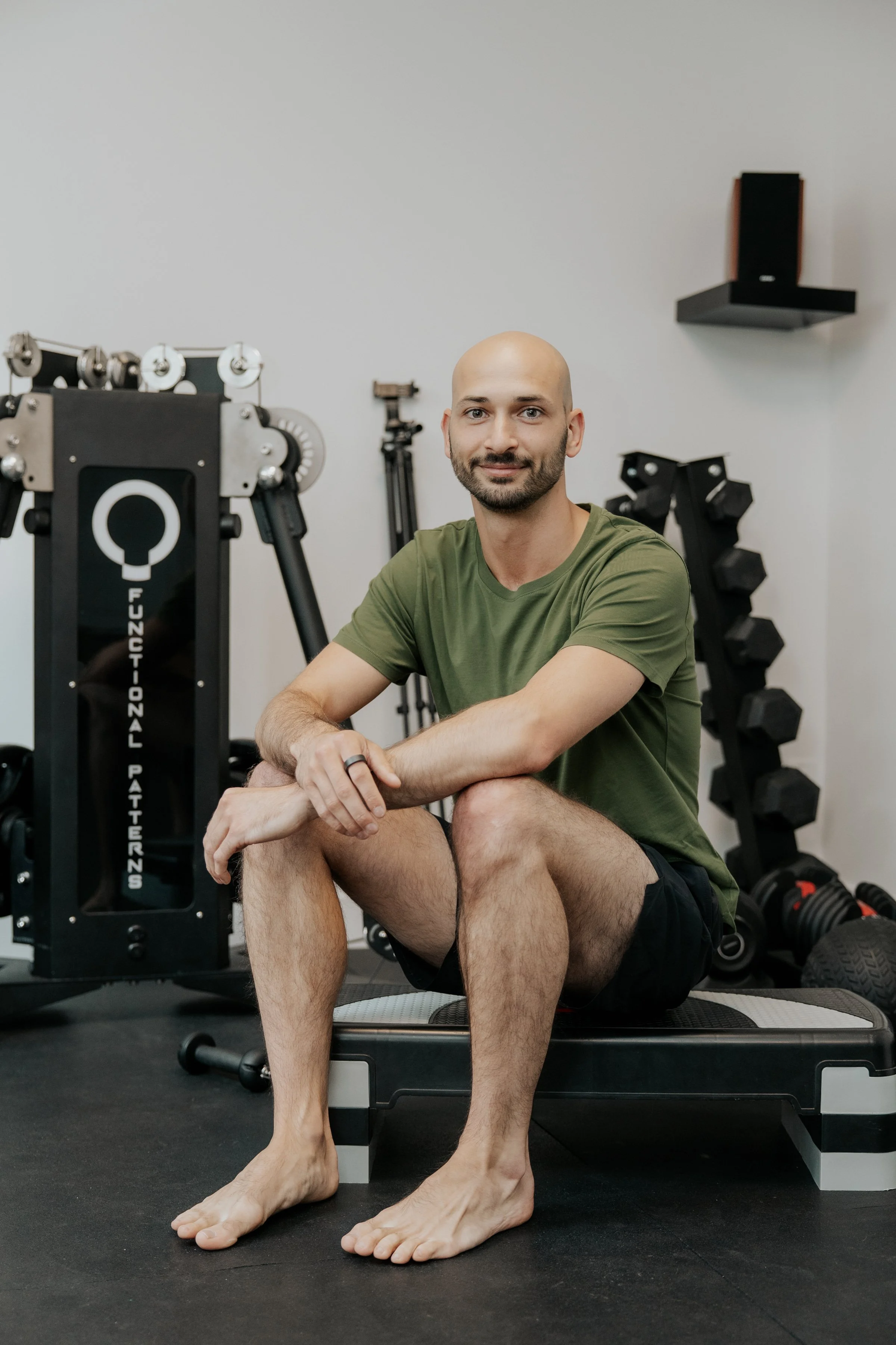 Un homme assis sur un banc de musculation dans une salle de sport, portant un t-shirt vert et des shorts noirs, avec des haltères et un équipement d'exercice en arrière-plan.