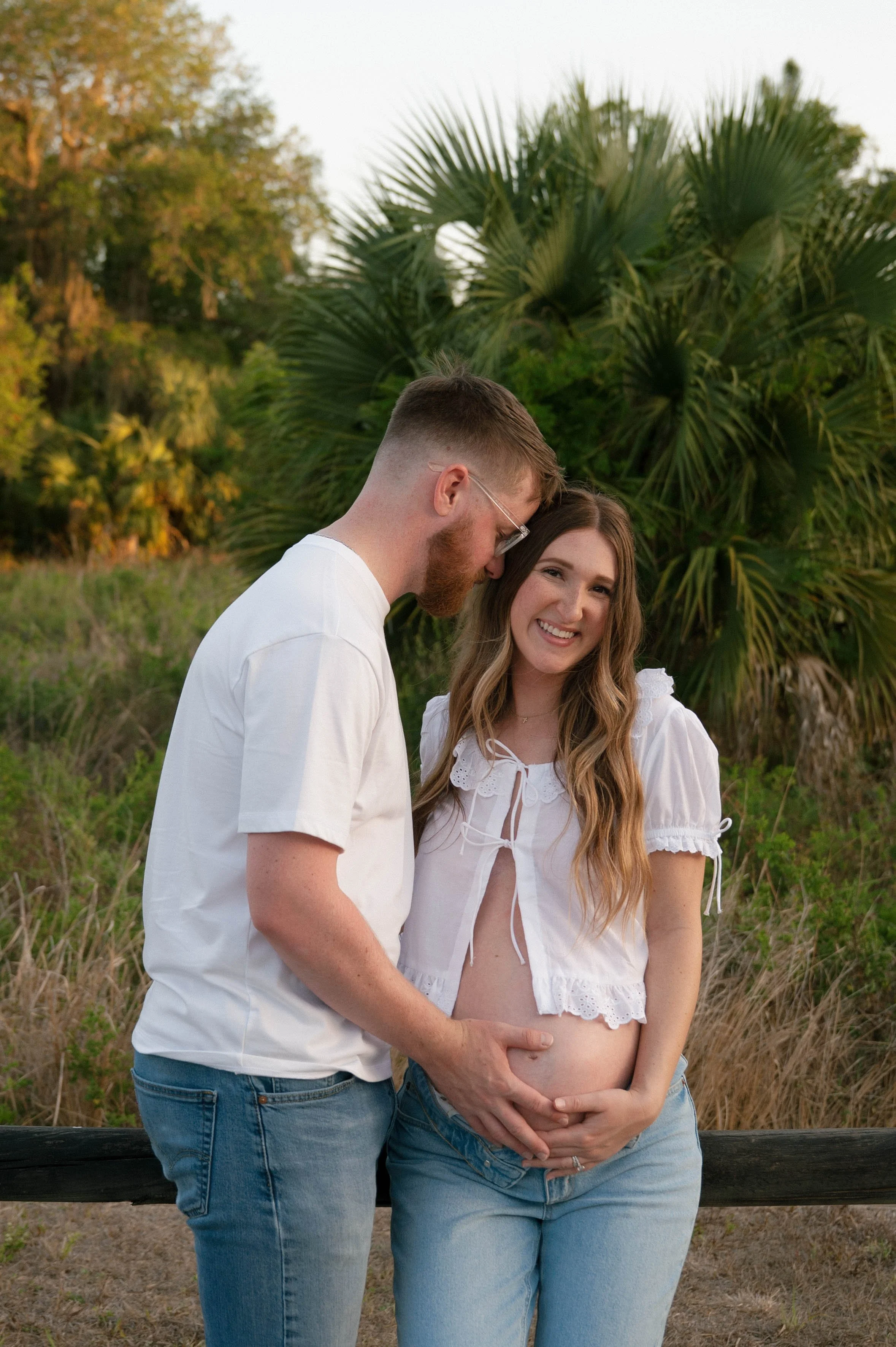 A pregnant woman and a man standing outside near a wooden fence, with greenery and tall plants in the background. The man is gently touching the woman's belly, and they are smiling and looking at the camera.