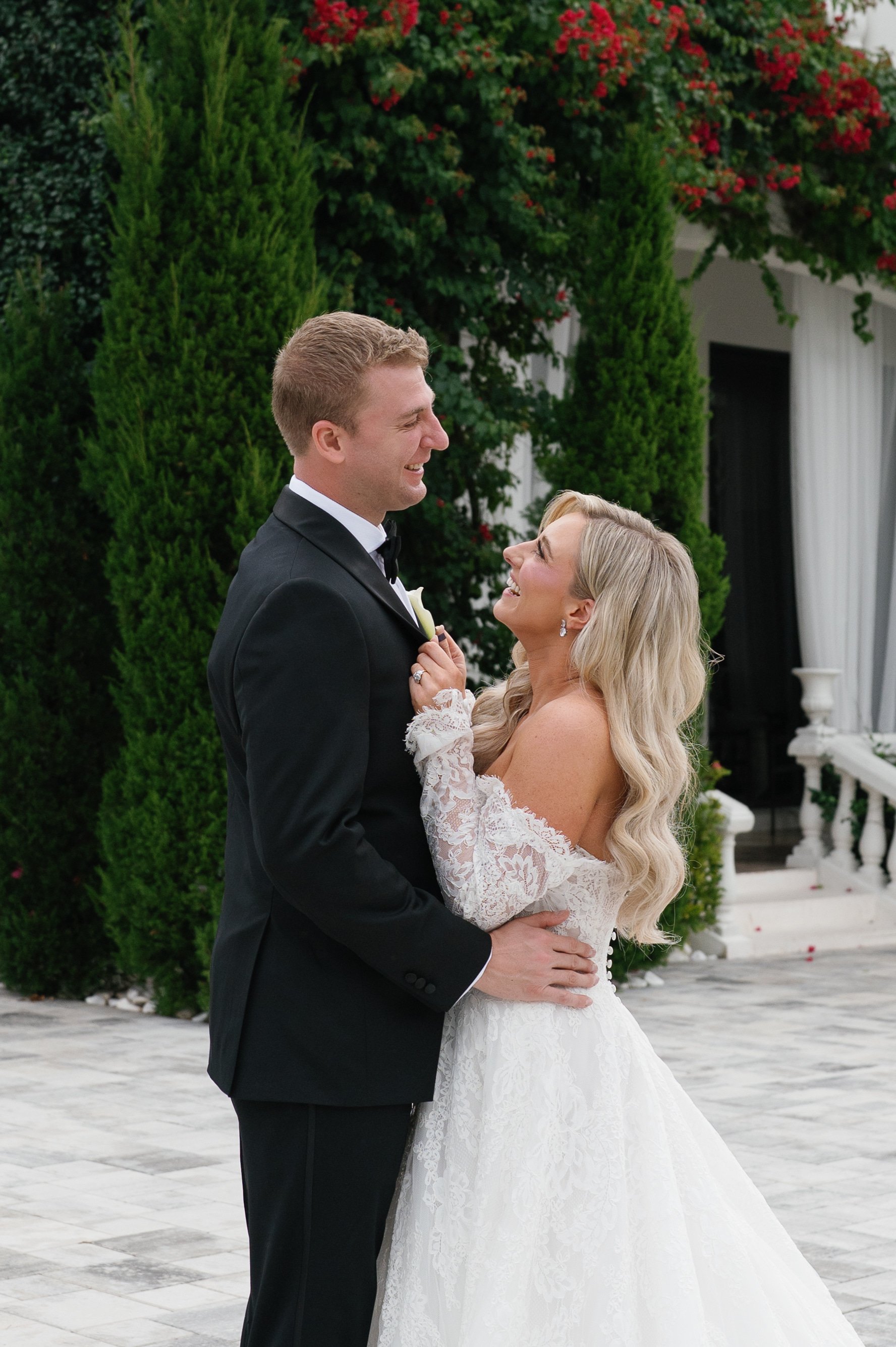 A newlywed couple, the groom in a black tuxedo and the bride in a white lace wedding gown, sharing a joyful moment outdoors with lush green trees and flowering bushes in the background.
