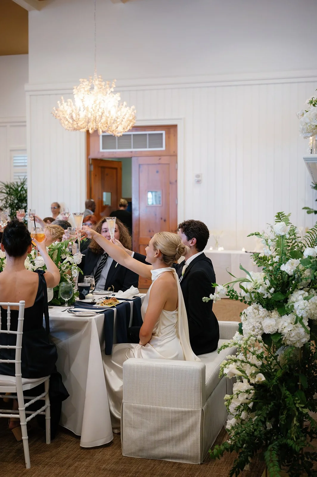 People celebrating at a wedding reception with champagne toasts, surrounded by floral arrangements in a decorated event space.