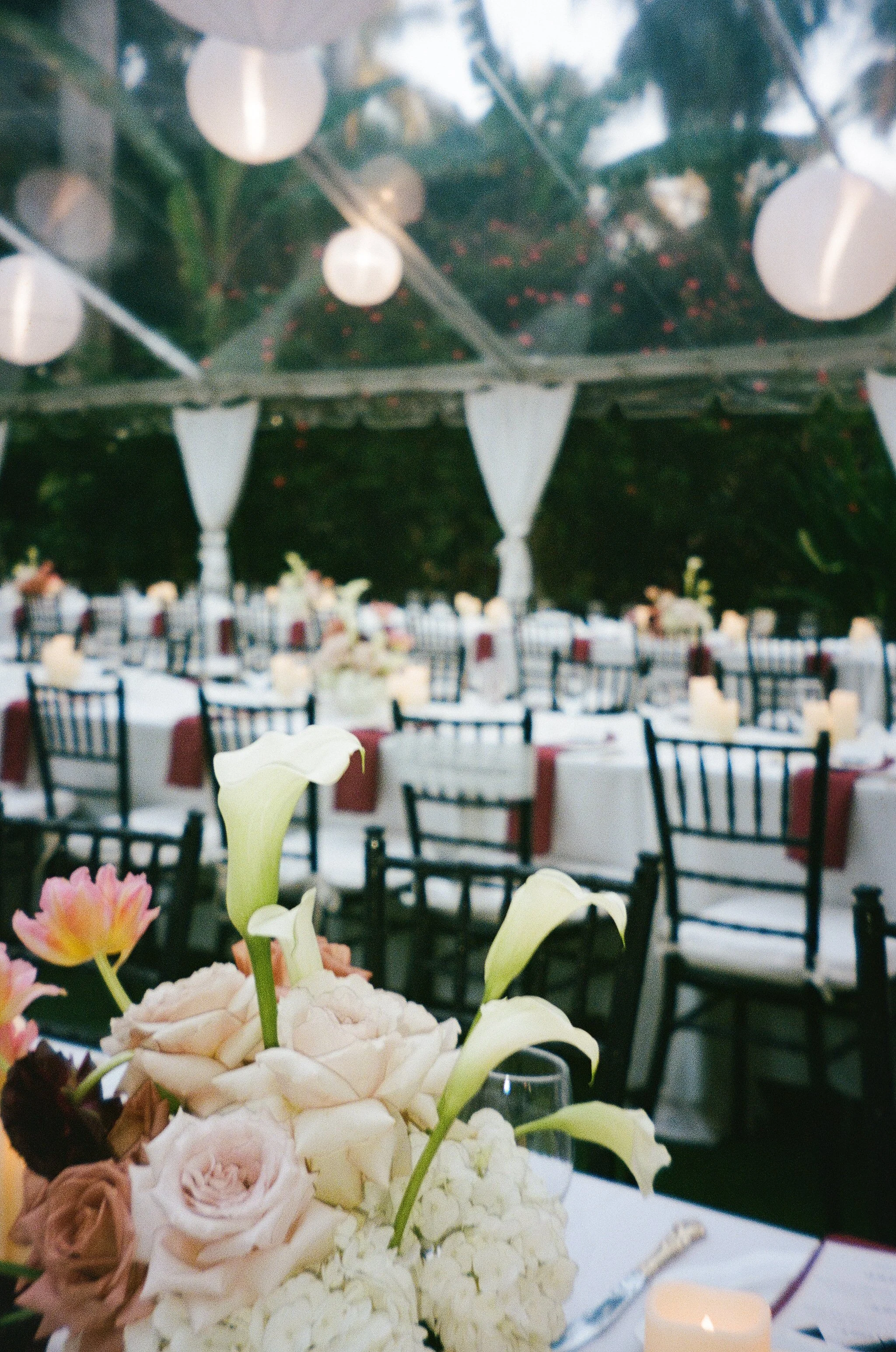 Close-up of floral arrangements including calla lilies, roses, and hydrangeas on a table at an event with lit candles and a decorated ceiling in the background.