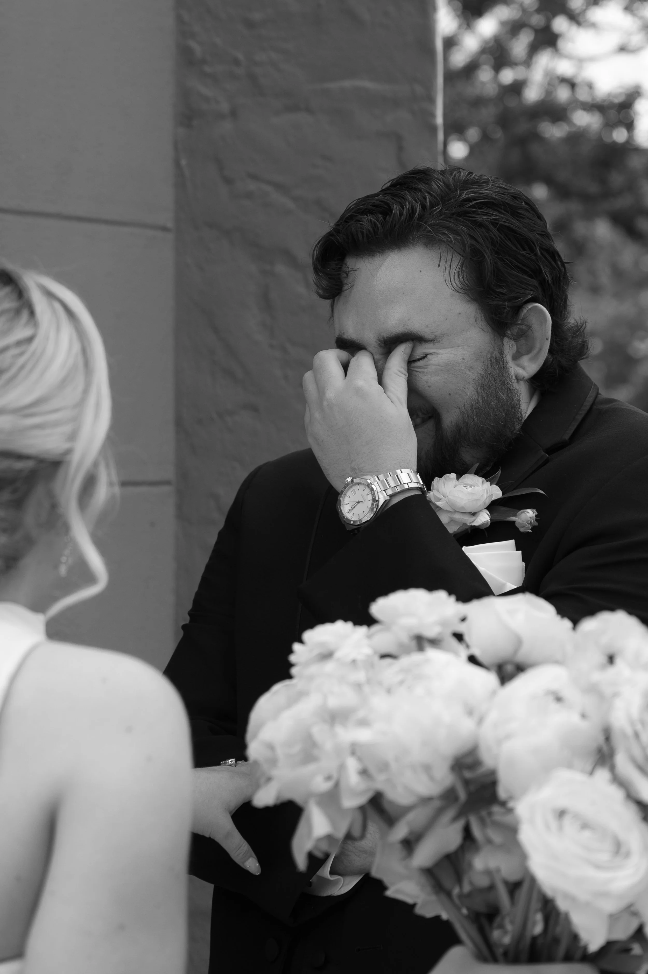 A man with dark wavy hair and a beard, wearing a suit and a watch, is holding his face with his hand and smiling with his eyes closed. He is sitting at a table with a bouquet of flowers in the foreground. A woman with light-colored hair, partly visib