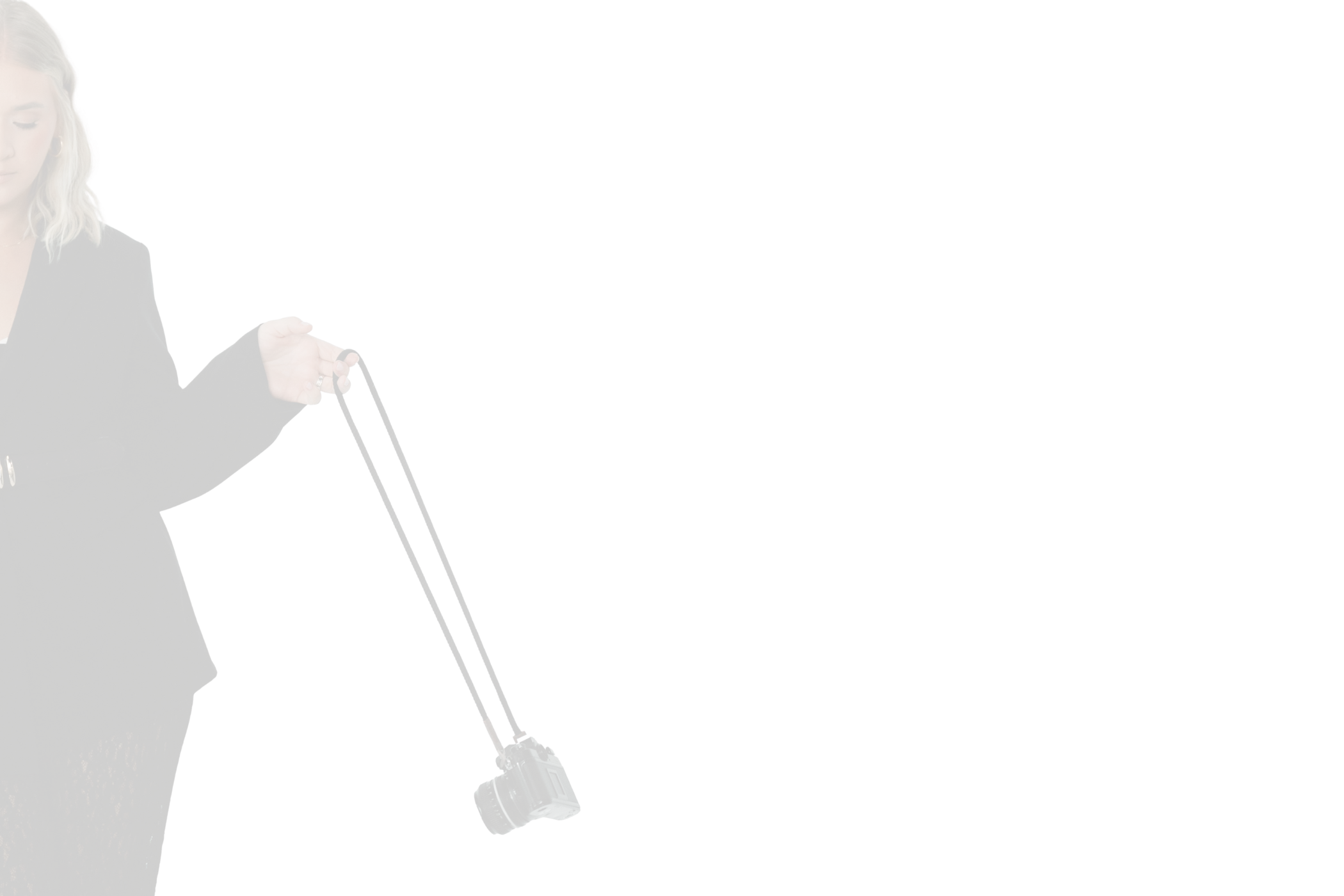 A woman with blonde hair in a black blazer holding a camera with a long lens, standing against a black background.