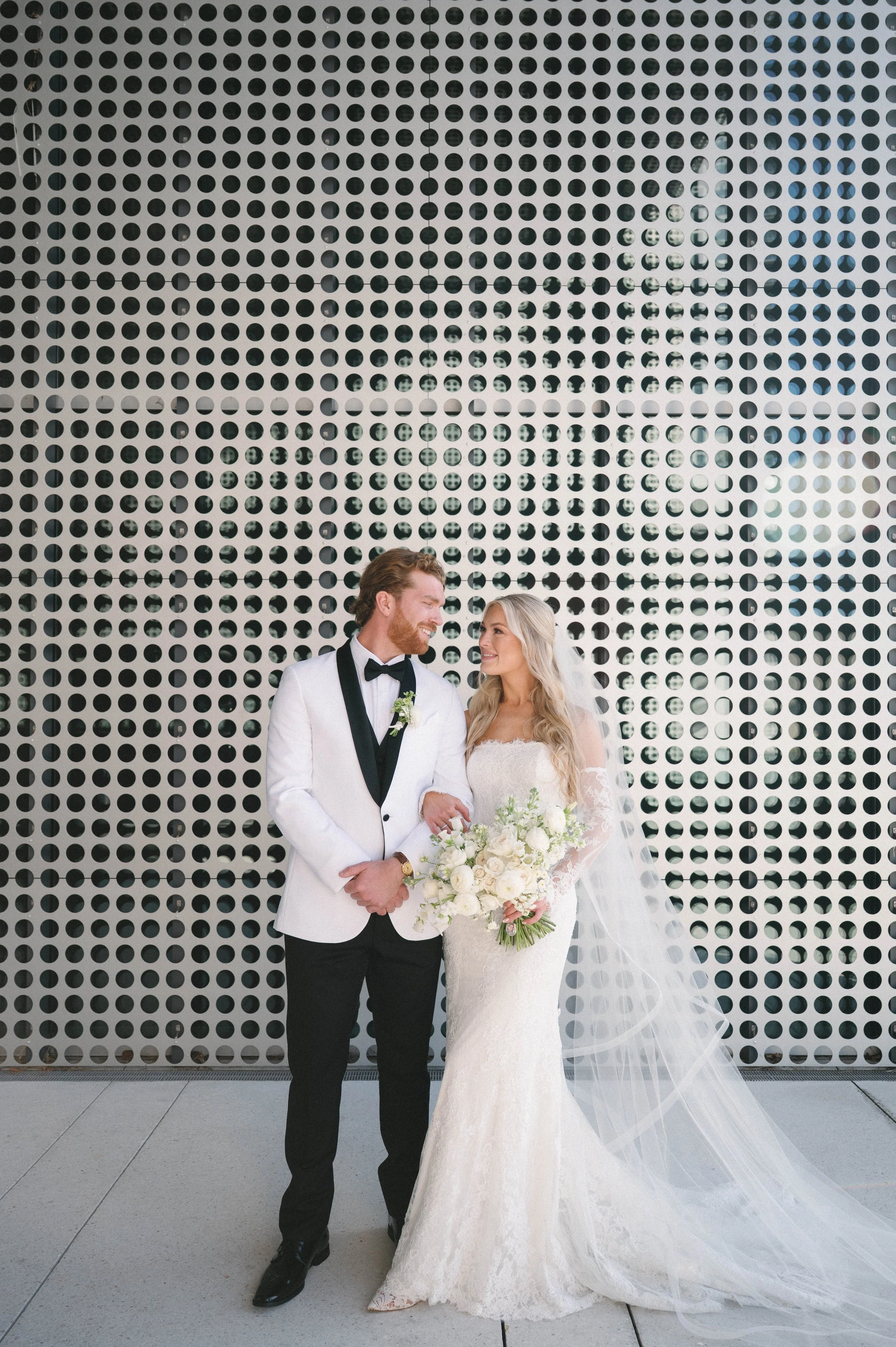 A bride and groom are standing together outside in front of a large decorative wall with circular cutouts. The groom is wearing a white tuxedo jacket with black lapels, black pants, and a bow tie. The bride is wearing a white lace wedding gown with l