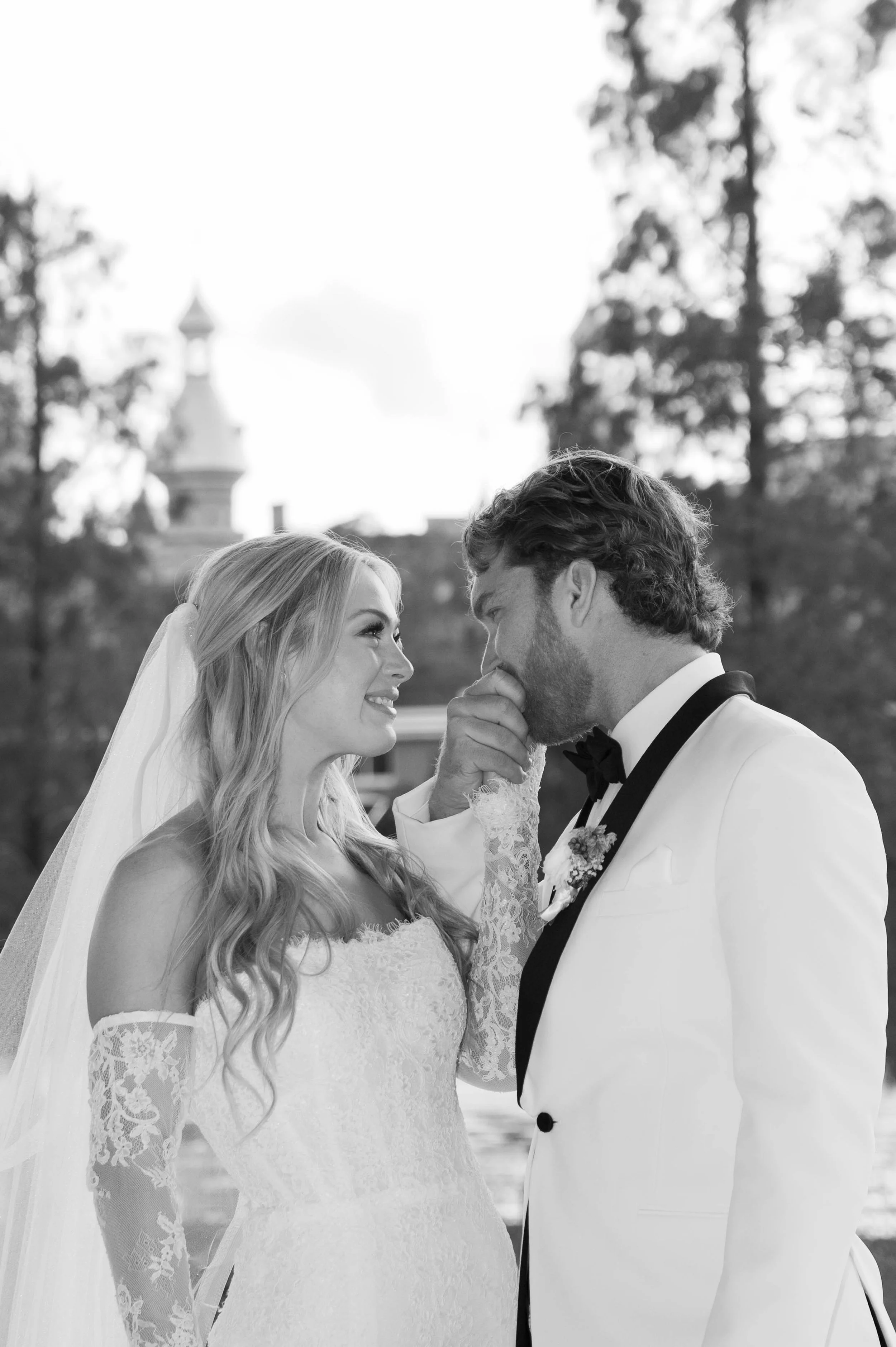 A black-and-white photograph of a bride and groom sharing a romantic moment outdoors, with trees and a blurred building in the background. The groom is kissing the bride's forehead, and they are gazing into each other's eyes.