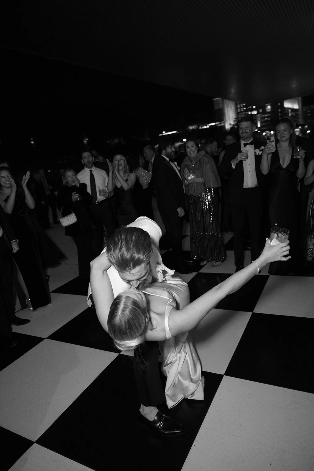A couple dancing closely on a checkered dance floor, surrounded by a group of party guests in formal attire smiling and holding drinks, at an elegant event.