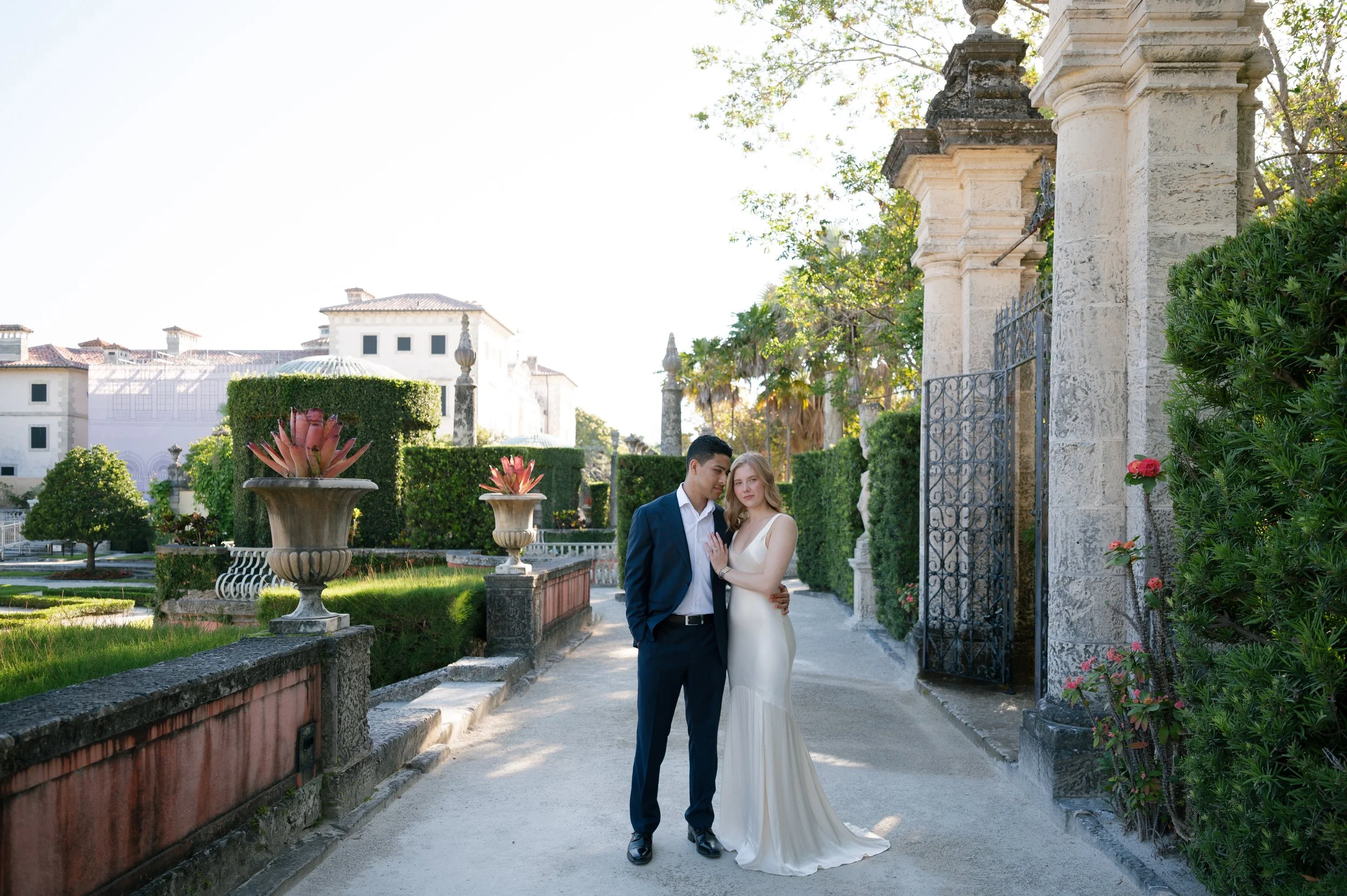 A young couple in formal attire standing close together in a lush garden with historic stone structures, decorative urns, and greenery during daylight.