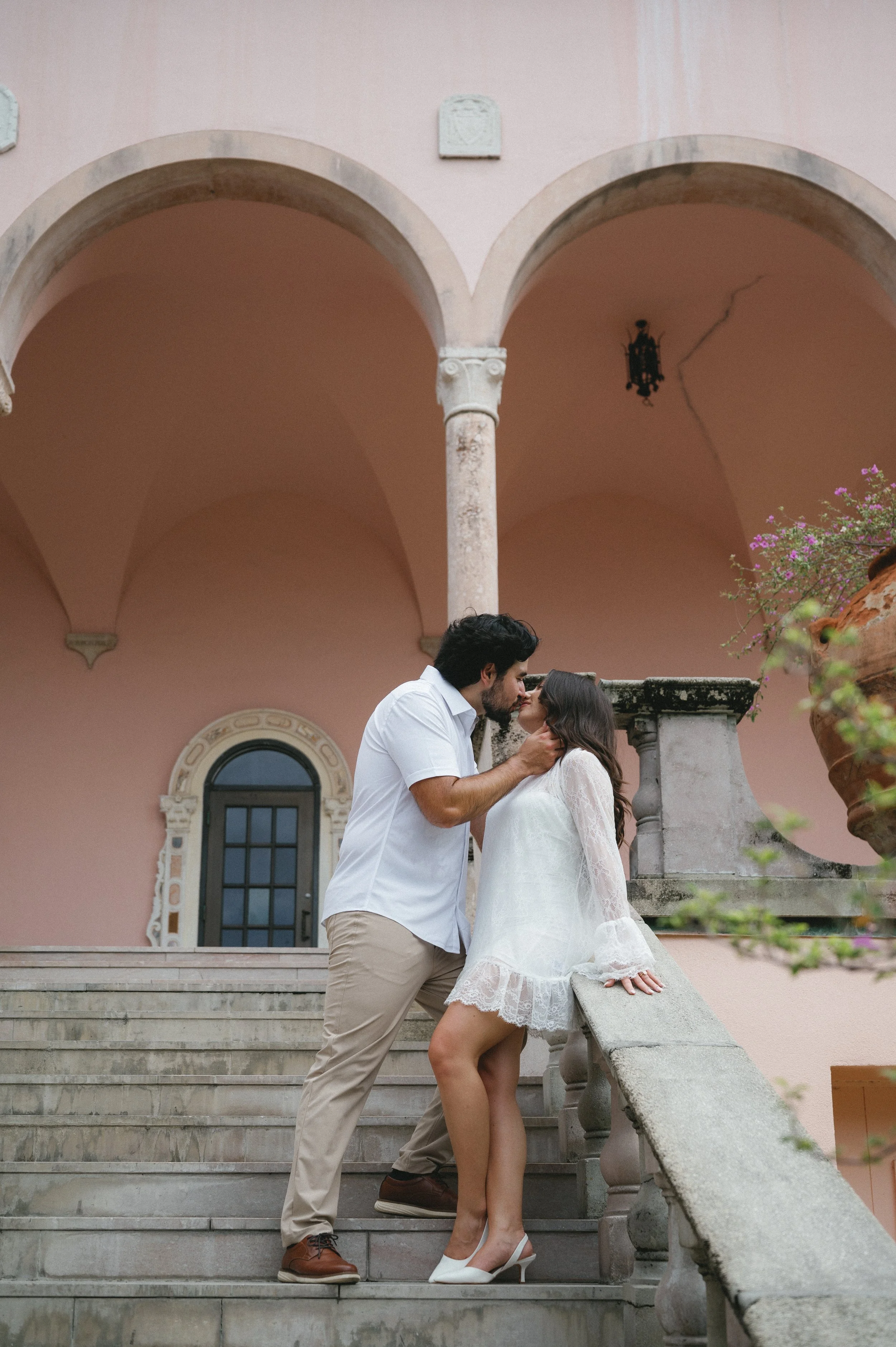 A man and woman sharing an intimate moment on a staircase in front of a pink building with arches and decorative columns. The woman is sitting on the steps, dressed in a white lace dress and heels, while the man stands close, holding her face and lea