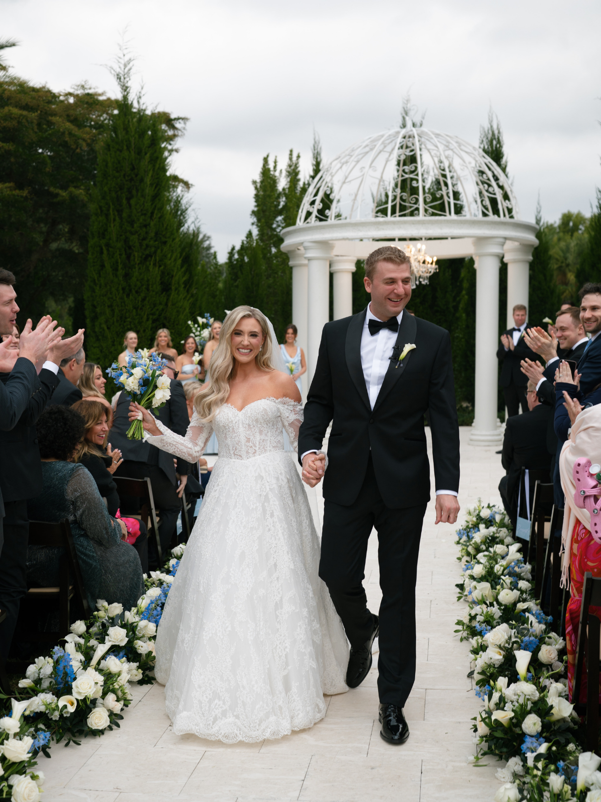 A bride and groom walking hand in hand down the aisle, smiling, at their outdoor wedding ceremony, surrounded by guests, flowers, and a white gazebo.