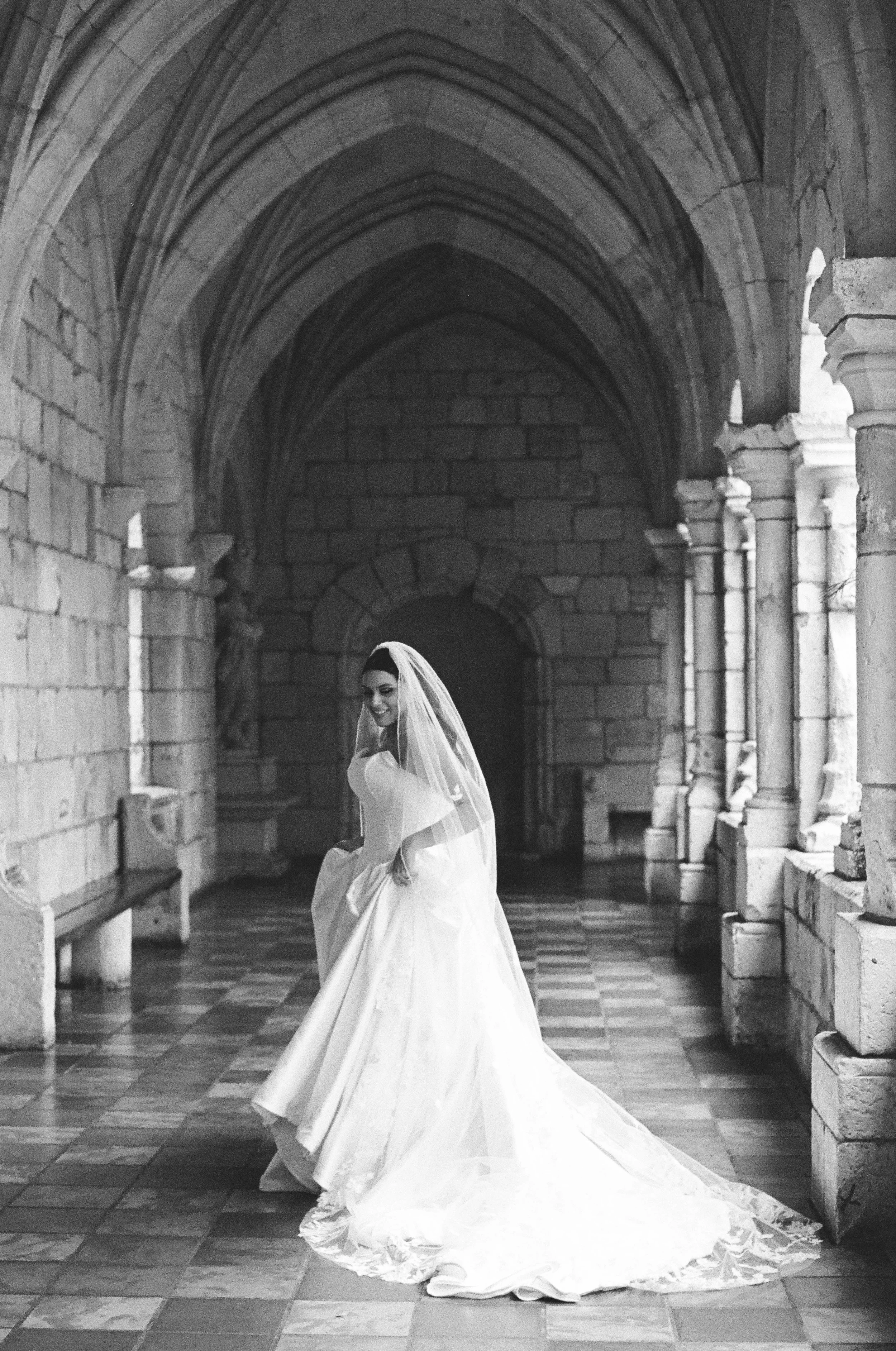 A woman in a wedding dress and veil standing in a Gothic-style stone hallway with arched ceiling, smiling and lifting her dress.