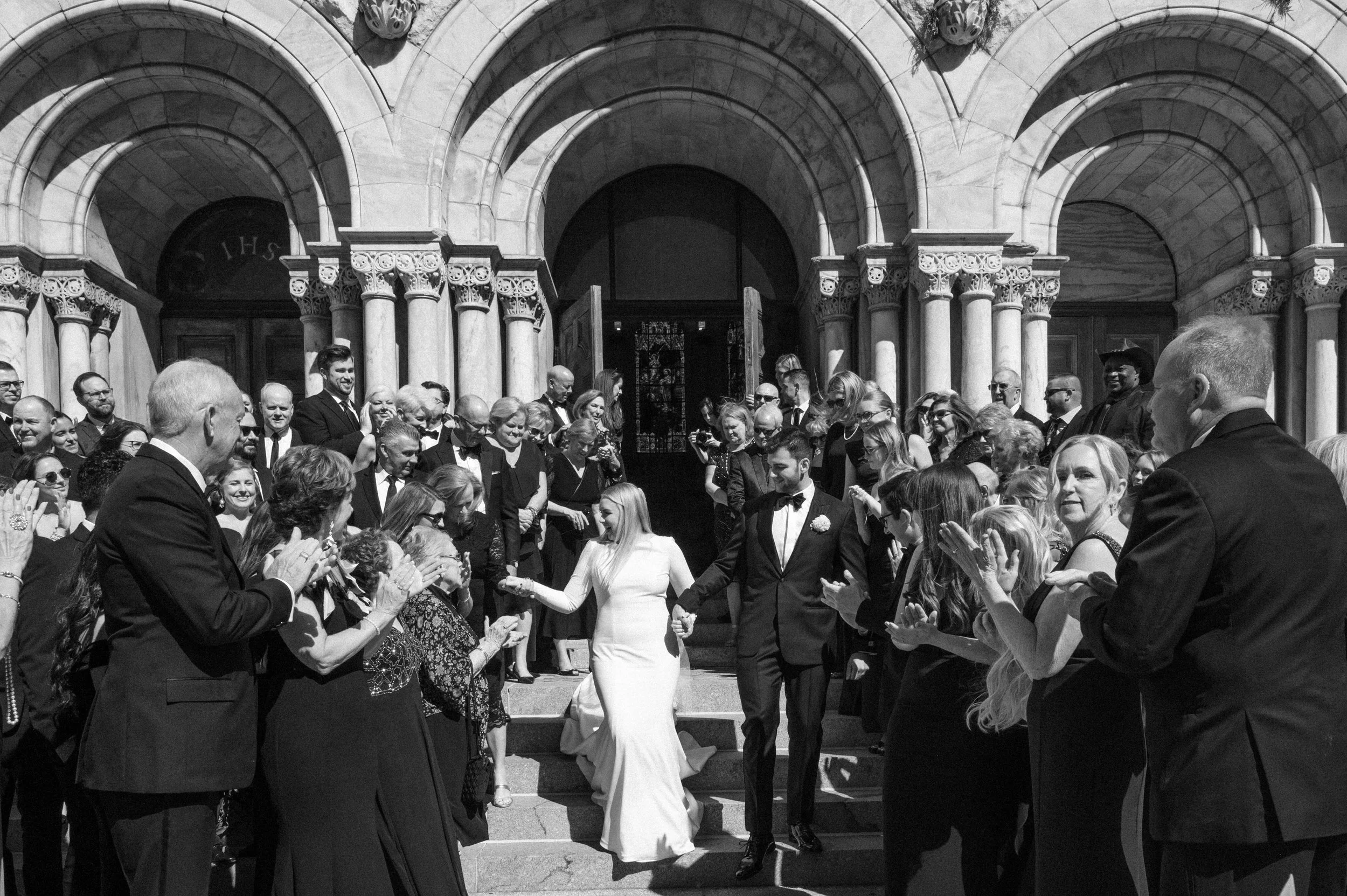A bride and groom holding hands at their wedding ceremony on the steps of a stone building, surrounded by guests.