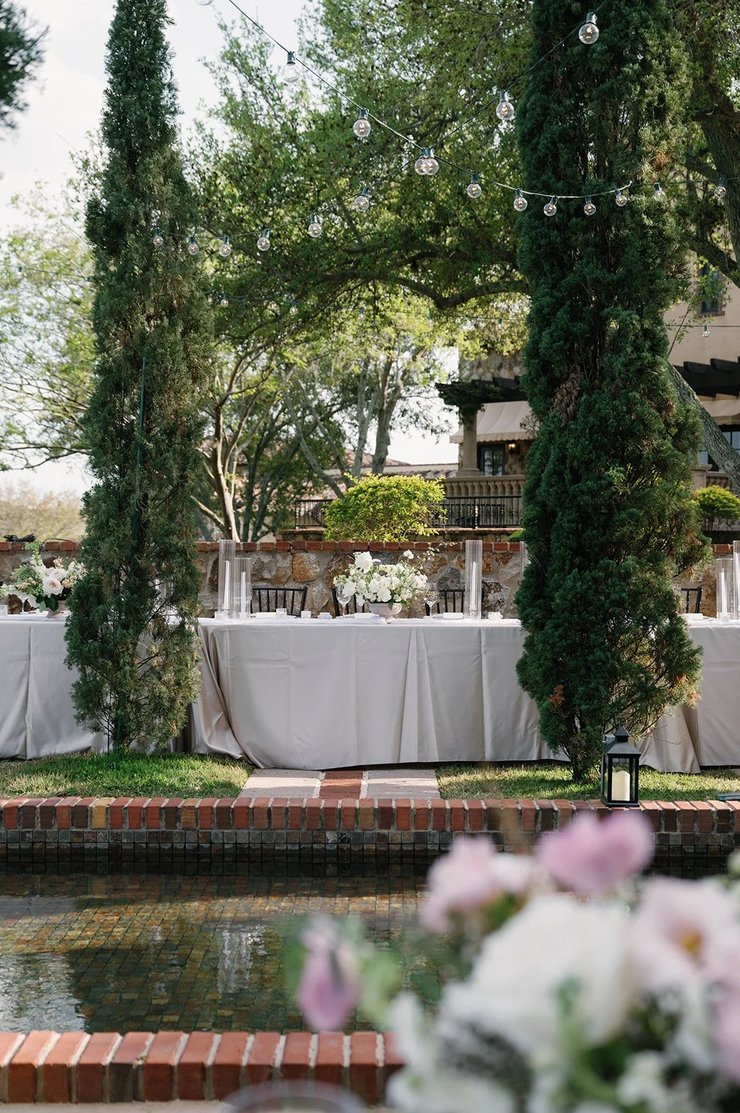 An outdoor event setup with a table draped in white cloth, decorated with white floral centerpieces, beneath a trellis with string lights surrounded by greenery and trees.